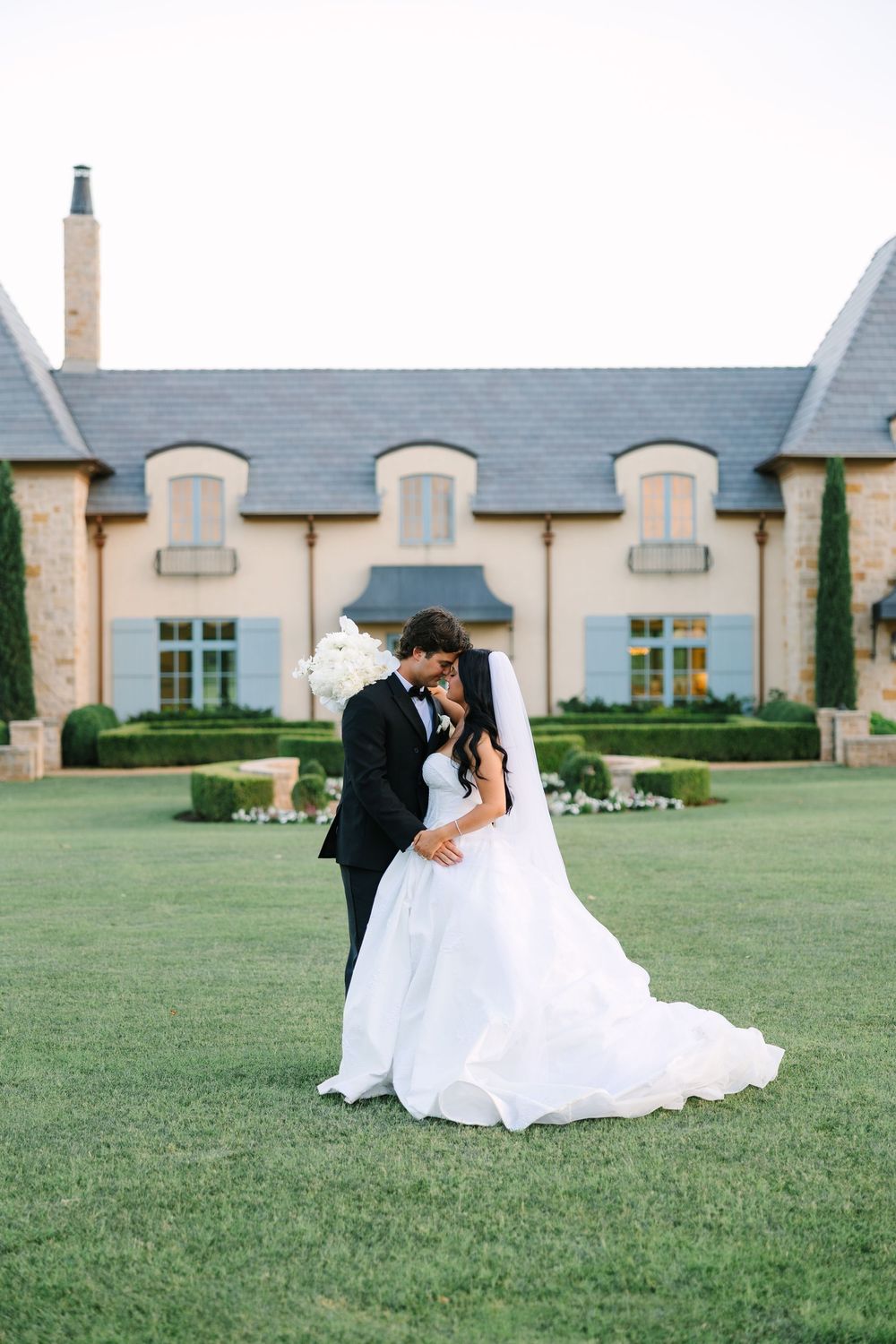 Bride and groom sharing a romantic kiss on lush green lawn in front of elegant French-style estate wedding venue.