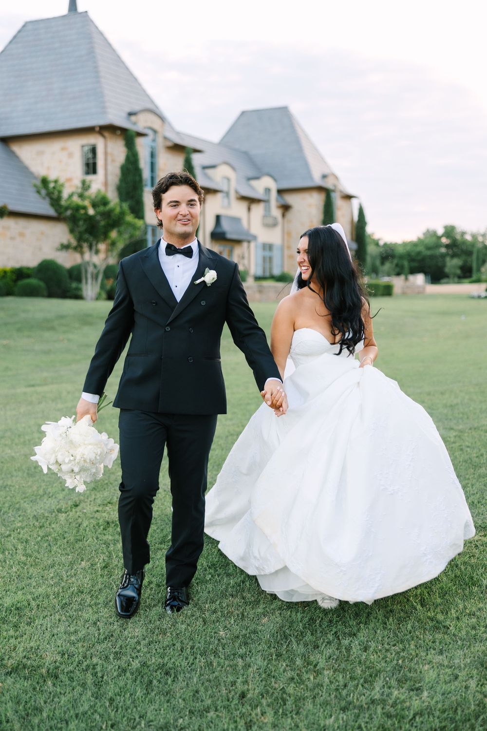 Bride and groom walk hand-in-hand on green lawn before elegant stone mansion, groom holding white bouquet.