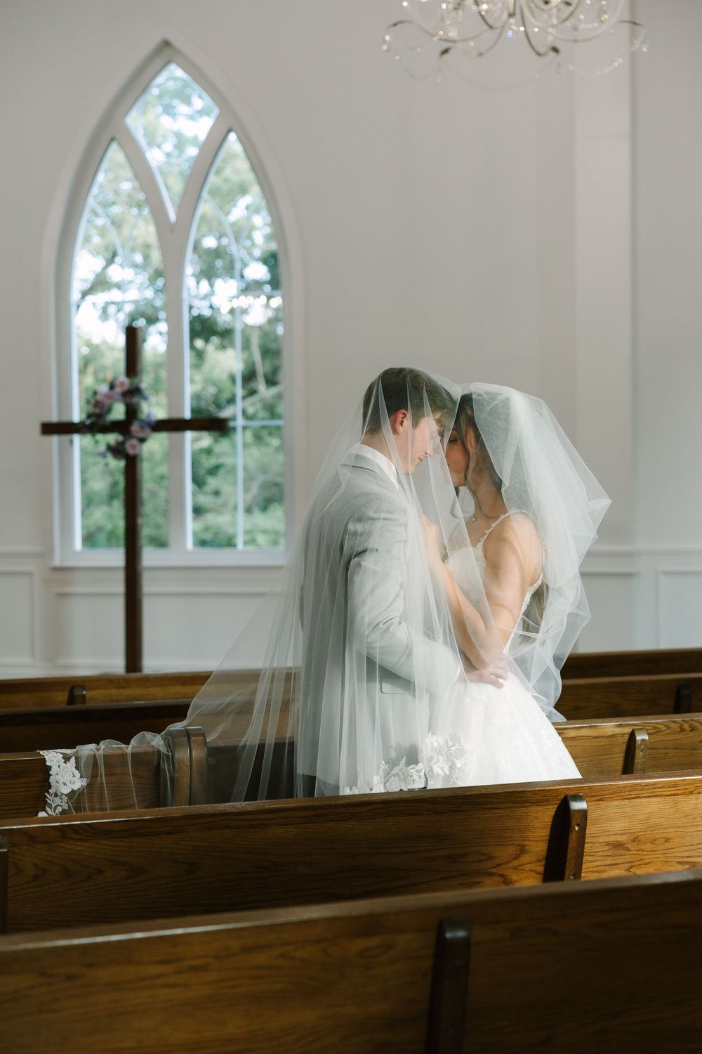 Bride and groom share a kiss under a veil in a white chapel with wooden pews and a Gothic arch window.