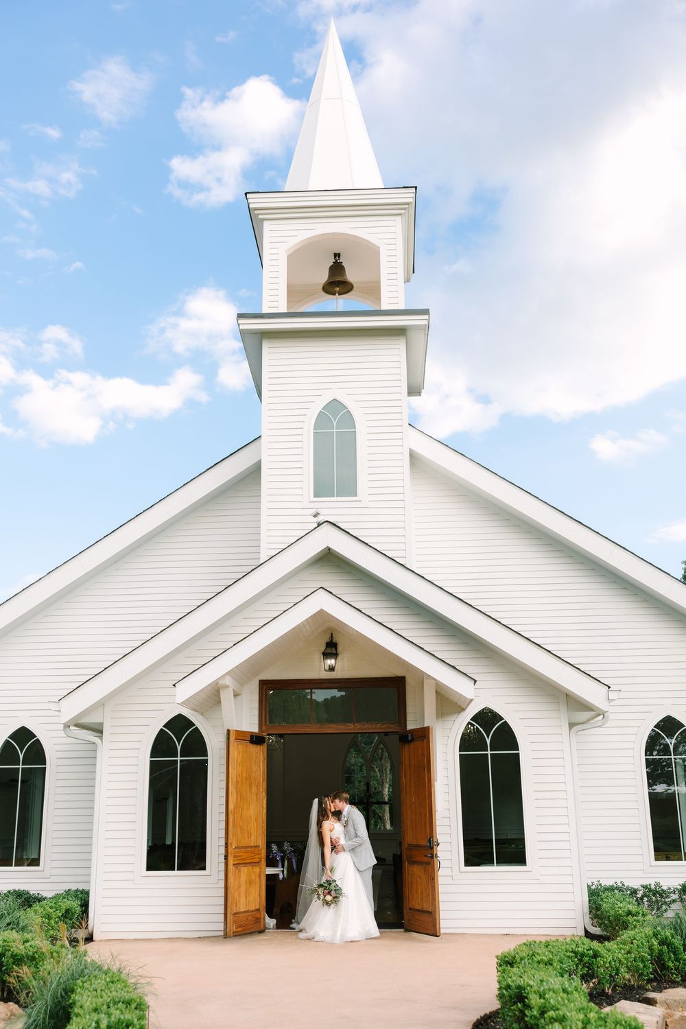 Bride and groom kissing at the entrance of a white chapel with a steeple on their wedding day.