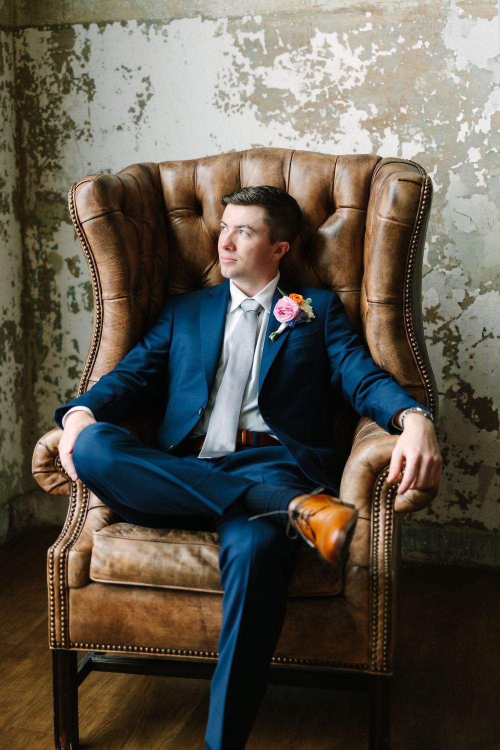 Groom in navy suit with floral boutonniere sits relaxed in brown leather wingback chair against rustic wall.