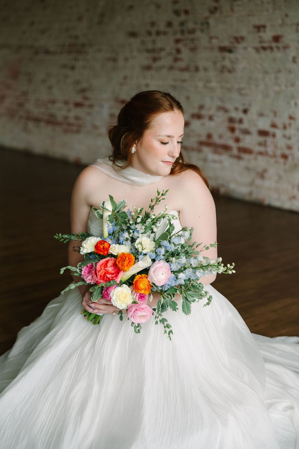 A bride in a white tulle gown holds a colorful bouquet of roses and greenery, seated before a rustic brick wall.