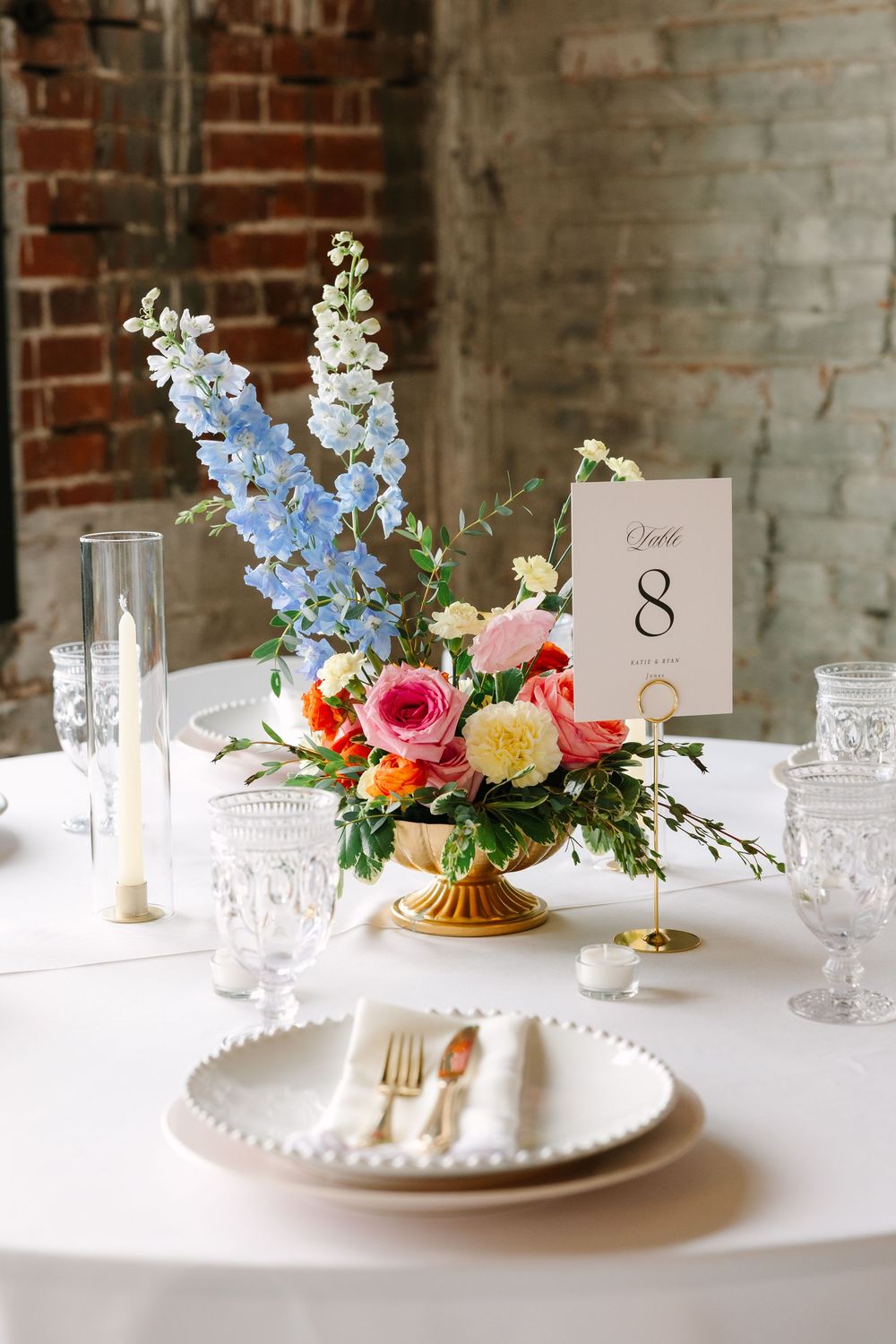Elegant wedding table with colorful floral centerpiece, crystal glasses, gold cutlery, and table number 8 on white linen.