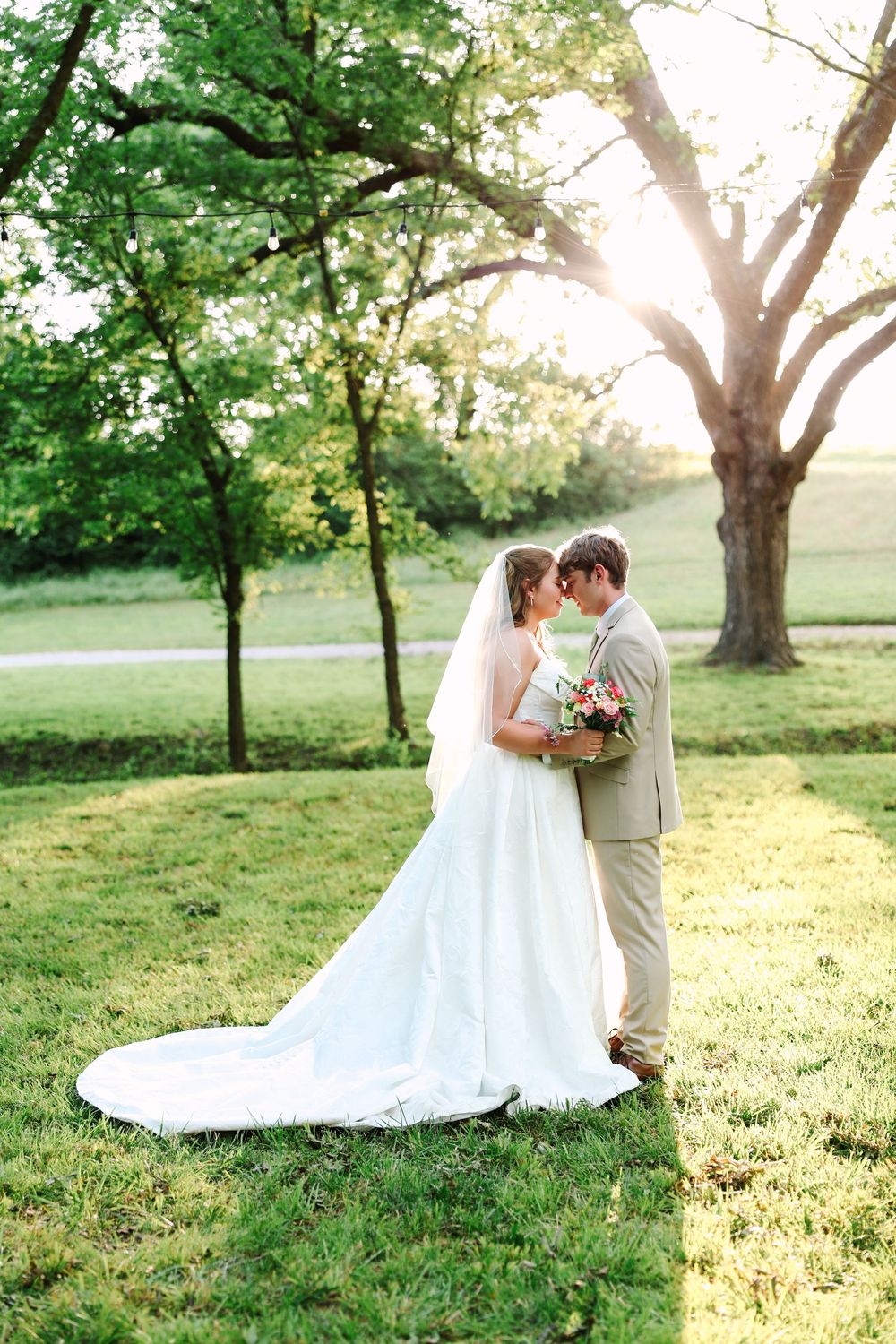 Bride and groom share a romantic moment outdoors at golden hour, surrounded by lush green trees on a sunlit lawn.