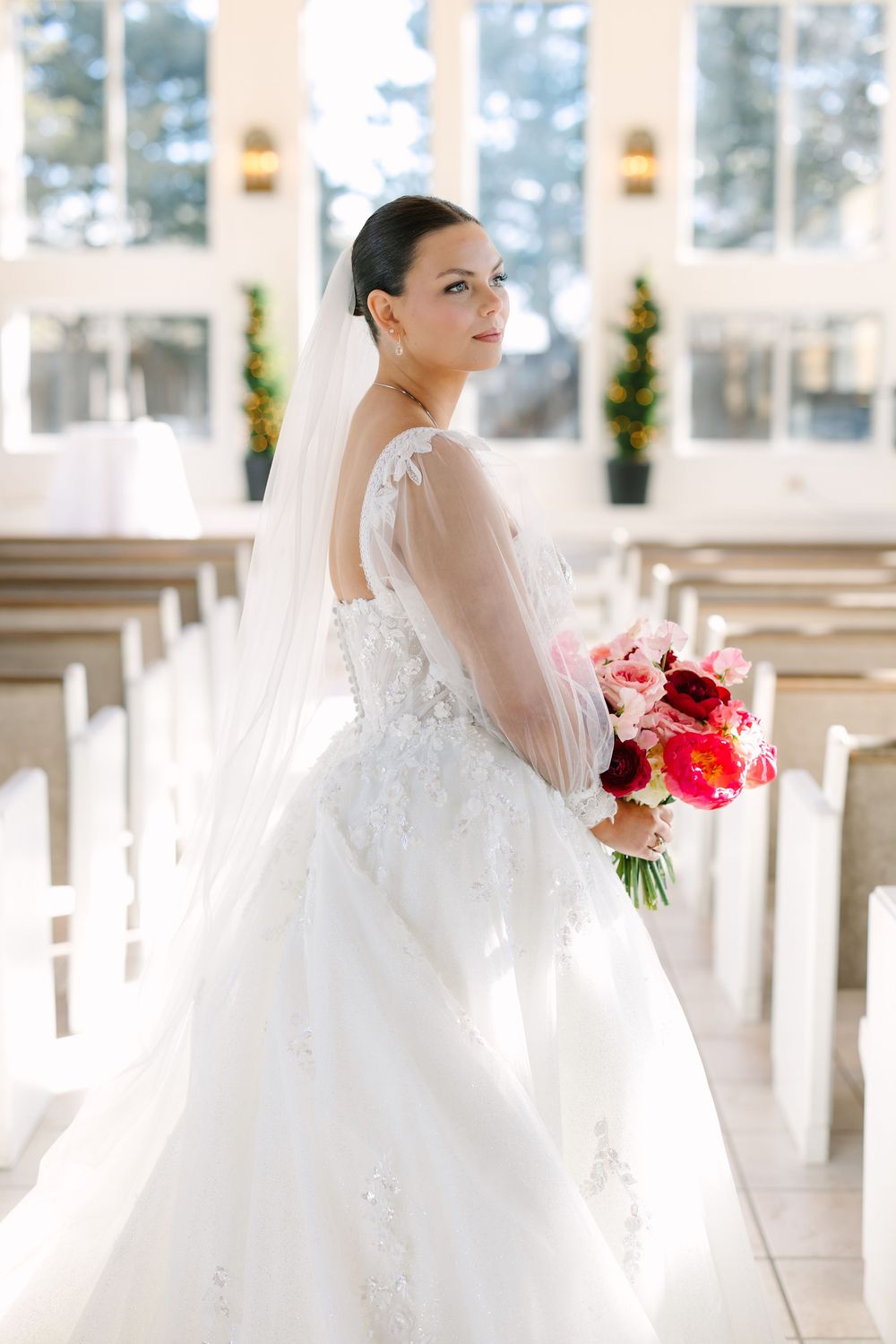 Elegant bride in a white lace ball gown and veil holding a pink and red bouquet inside a bright white chapel.