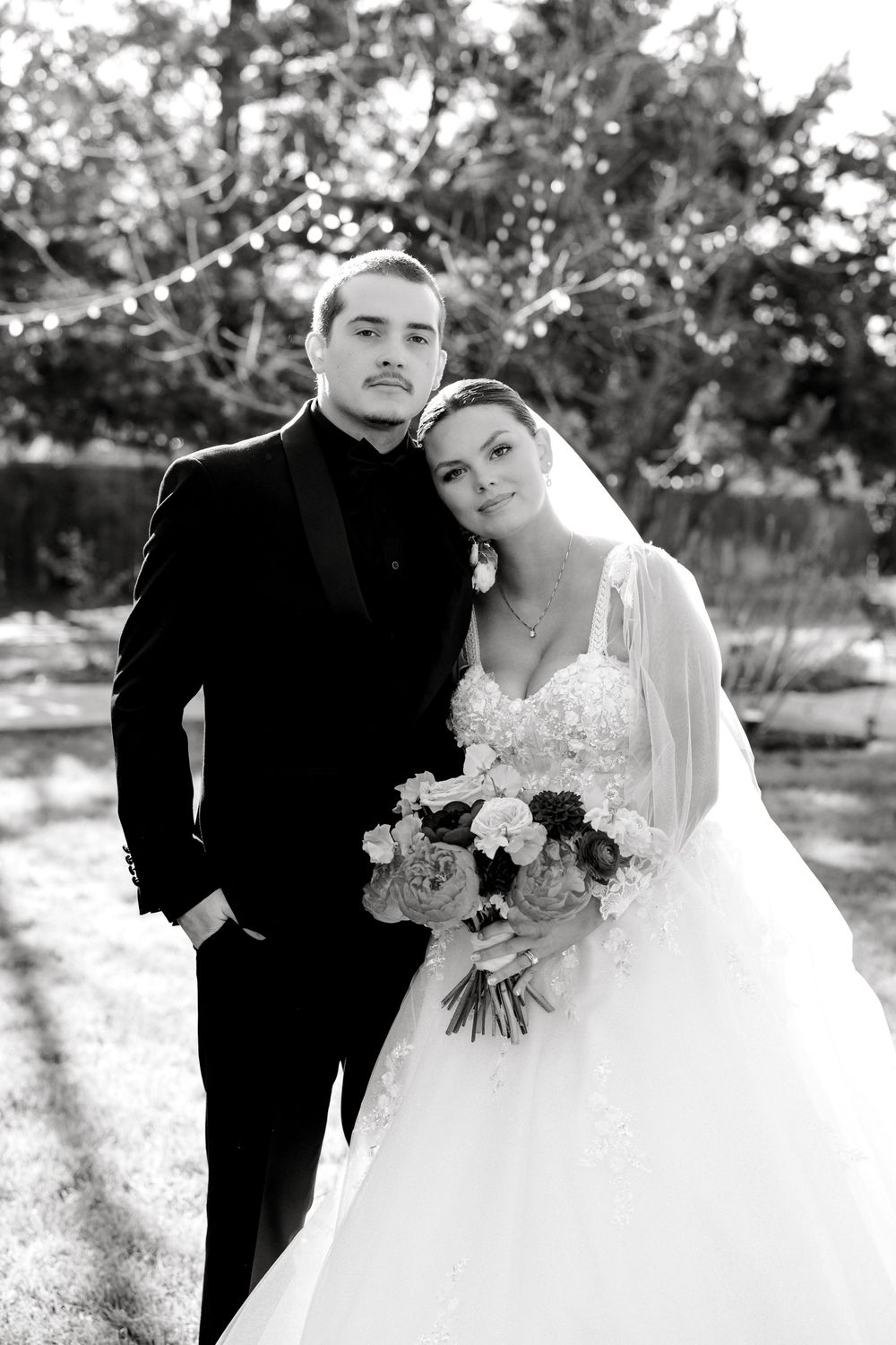 Bride and groom pose together outdoors in black and white portrait, bride holding bouquet in wedding attire.