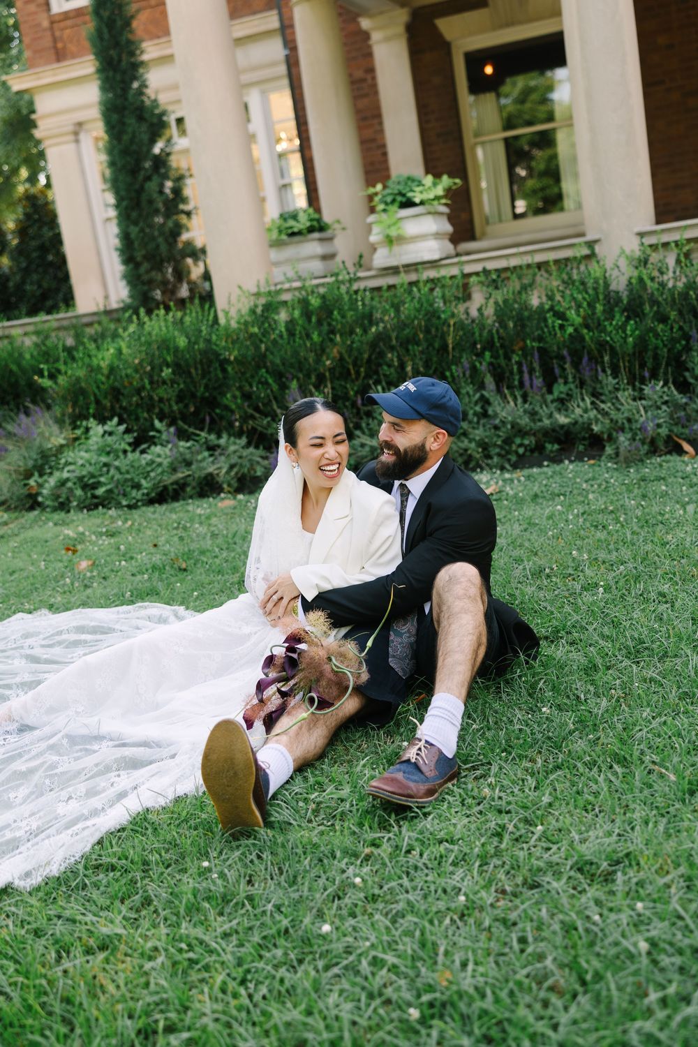 Bride and groom sitting on lush green grass laughing together, bride in white blazer with veil, groom in suit and cap.