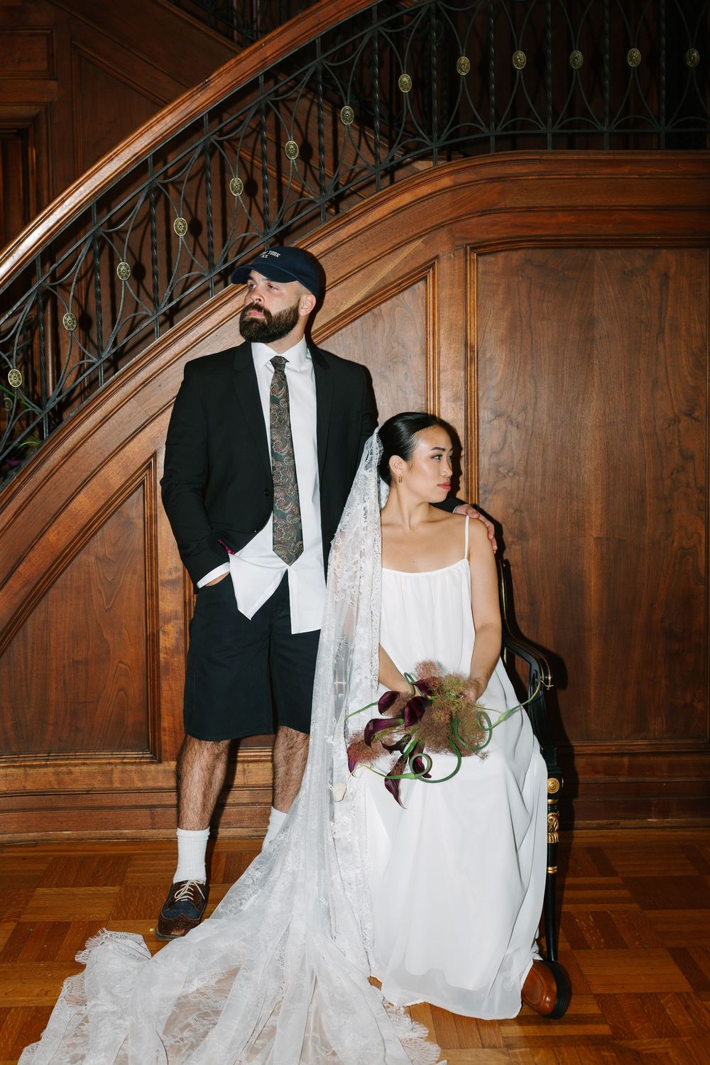 Wedding couple poses by ornate wooden staircase, bride in white gown with veil, groom in black blazer and shorts.