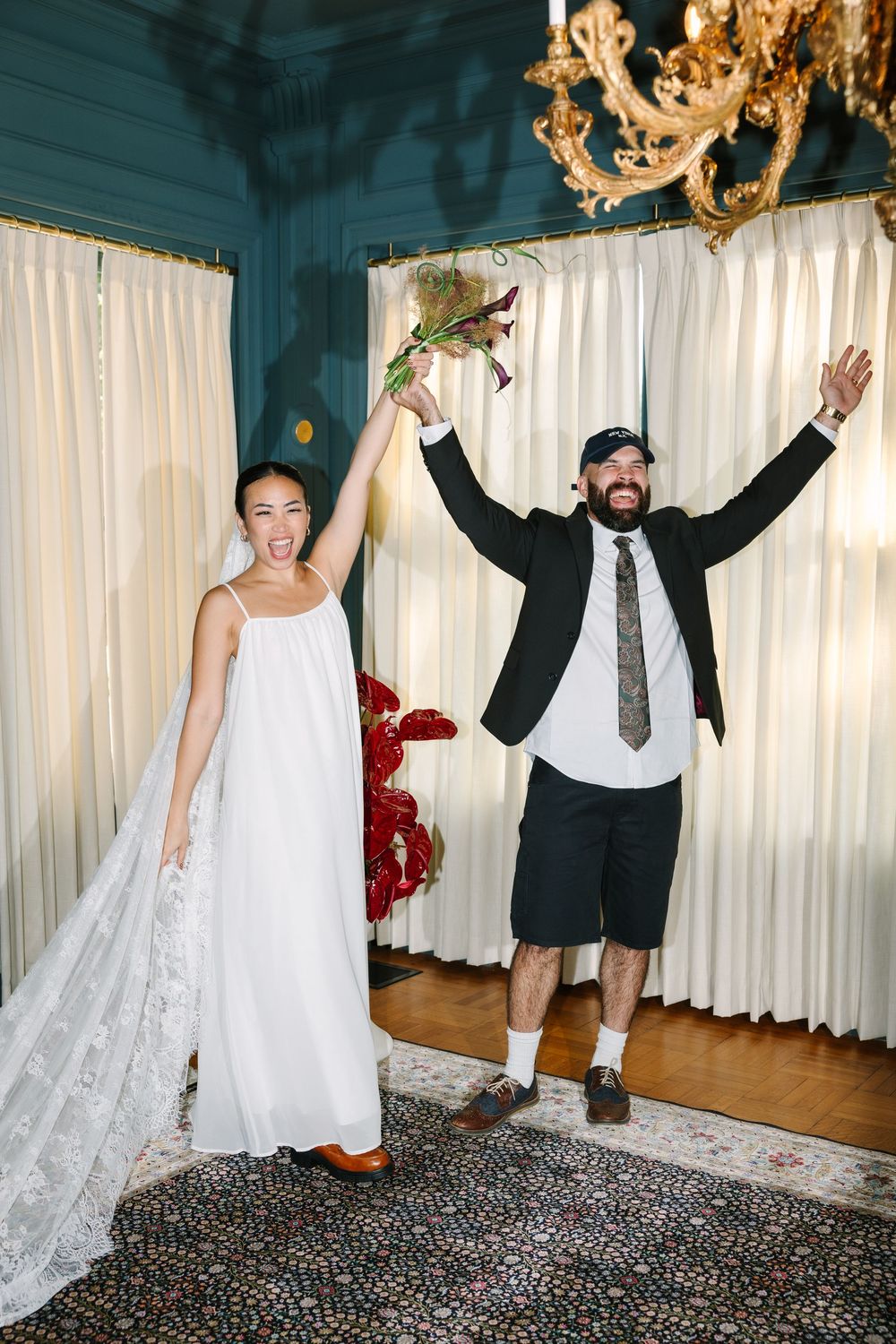 Joyful newlyweds celebrate their wedding, bride in white gown holding bouquet, groom in shorts and blazer raising arms.