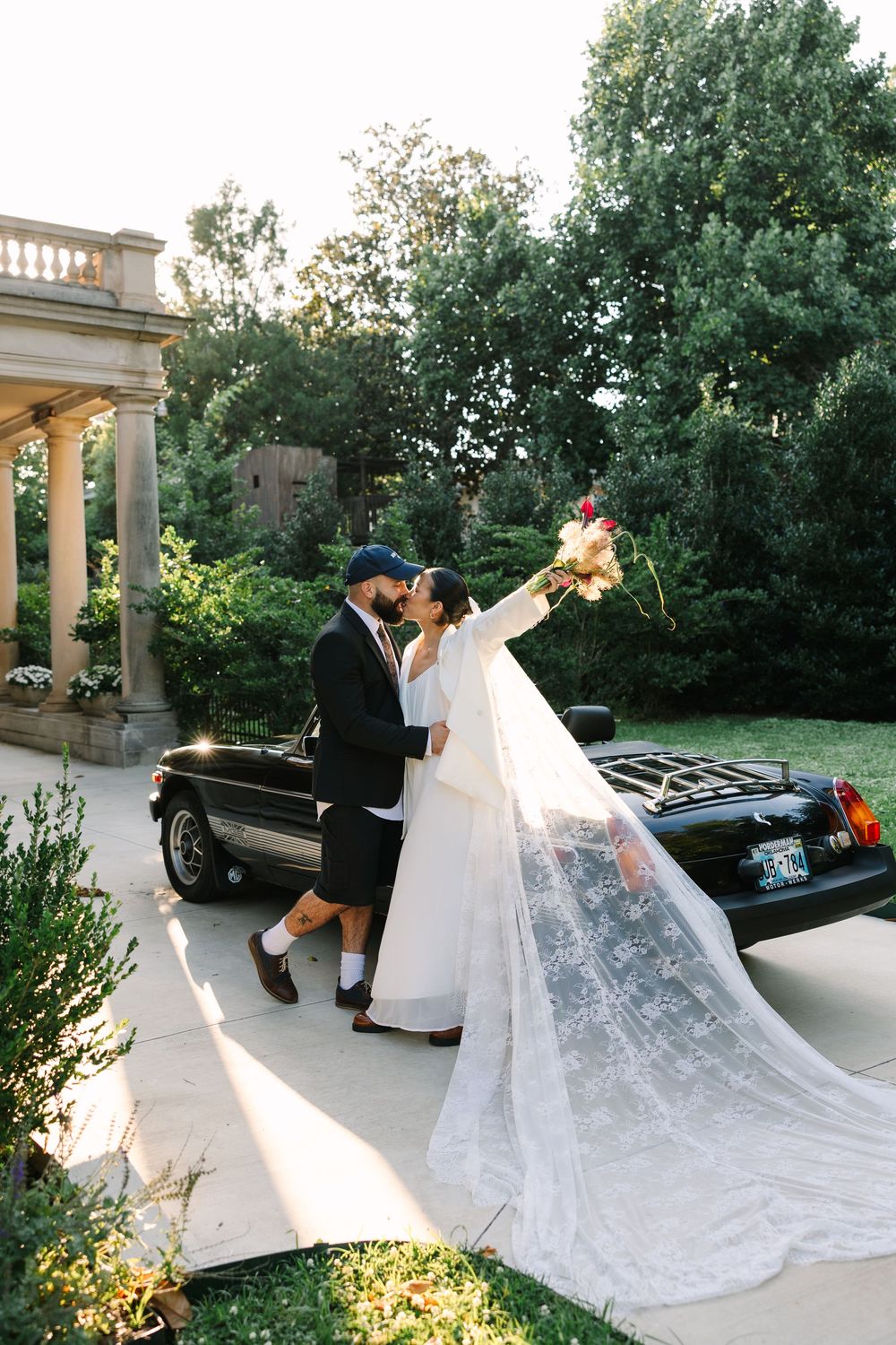Bride in lace veil raises bouquet while kissing groom in cap beside vintage black car in garden.