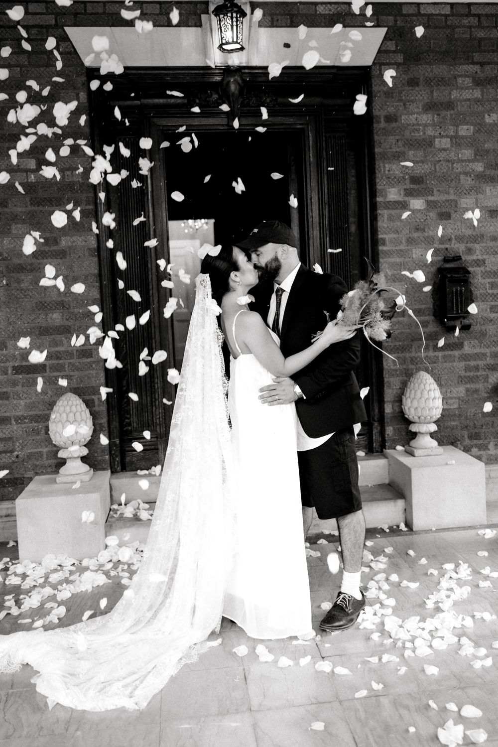 Black and white photo of a bride and groom kissing as flower petals rain down outside a brick church.
