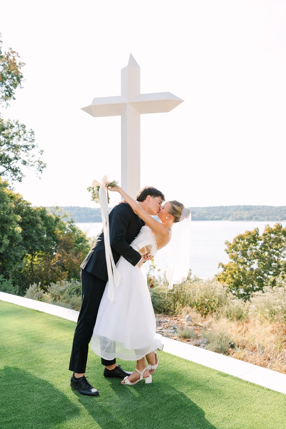 Bride and groom share a romantic dip kiss in front of a white cross with scenic waterfront views at outdoor wedding.