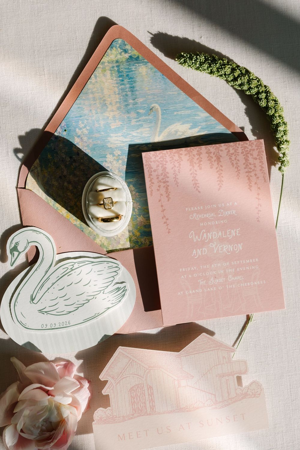 Elegant pink wedding invitation suite with swan motifs, floral envelope liner, rings, and greenery on white linen background.