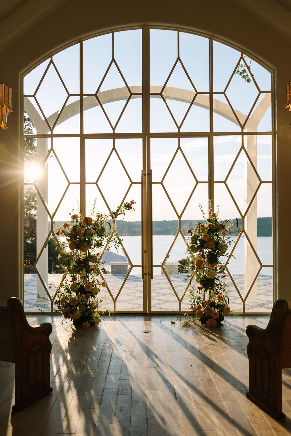 Elegant chapel with arched geometric windows overlooking a lake, flanked by floral arrangements in warm sunset light.