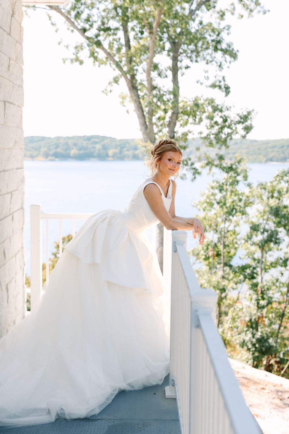 Bride in a white ballgown leans on a railing of a lakeside balcony surrounded by lush green trees.
