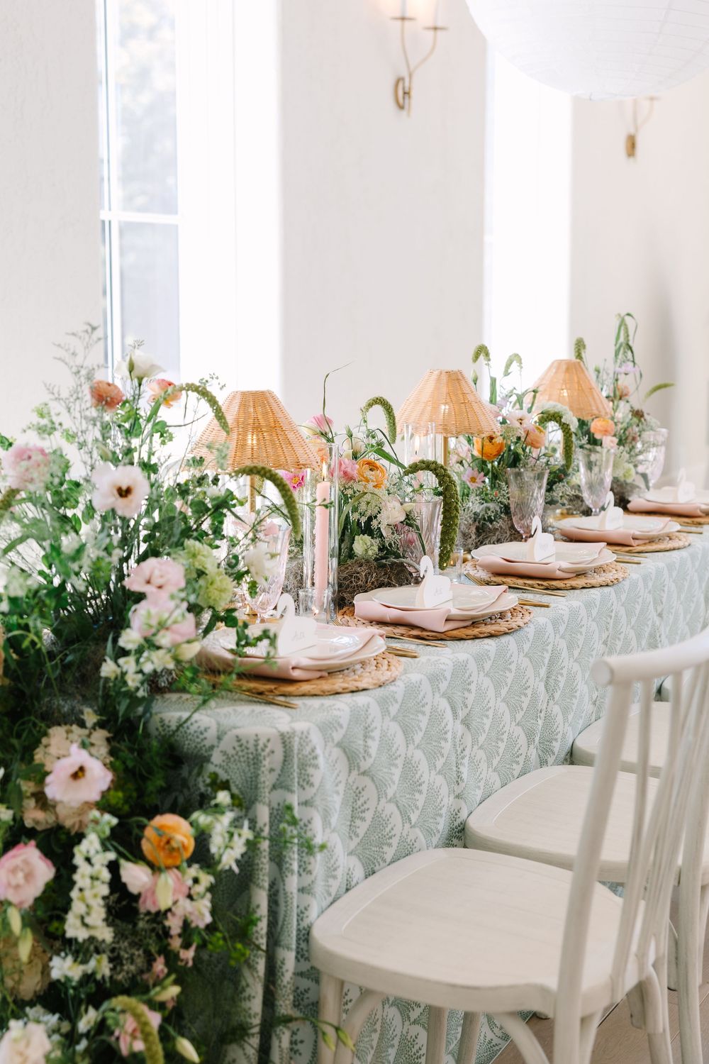 Elegant wedding reception table with floral runner, rattan lamps, pink candles, and sage green tablecloth in bright white room.