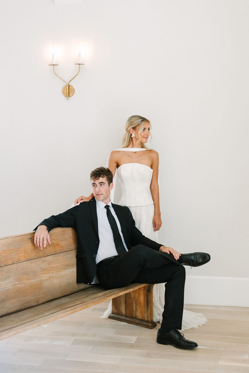 Bride in strapless white gown stands behind groom in black suit seated on wooden bench against white wall.