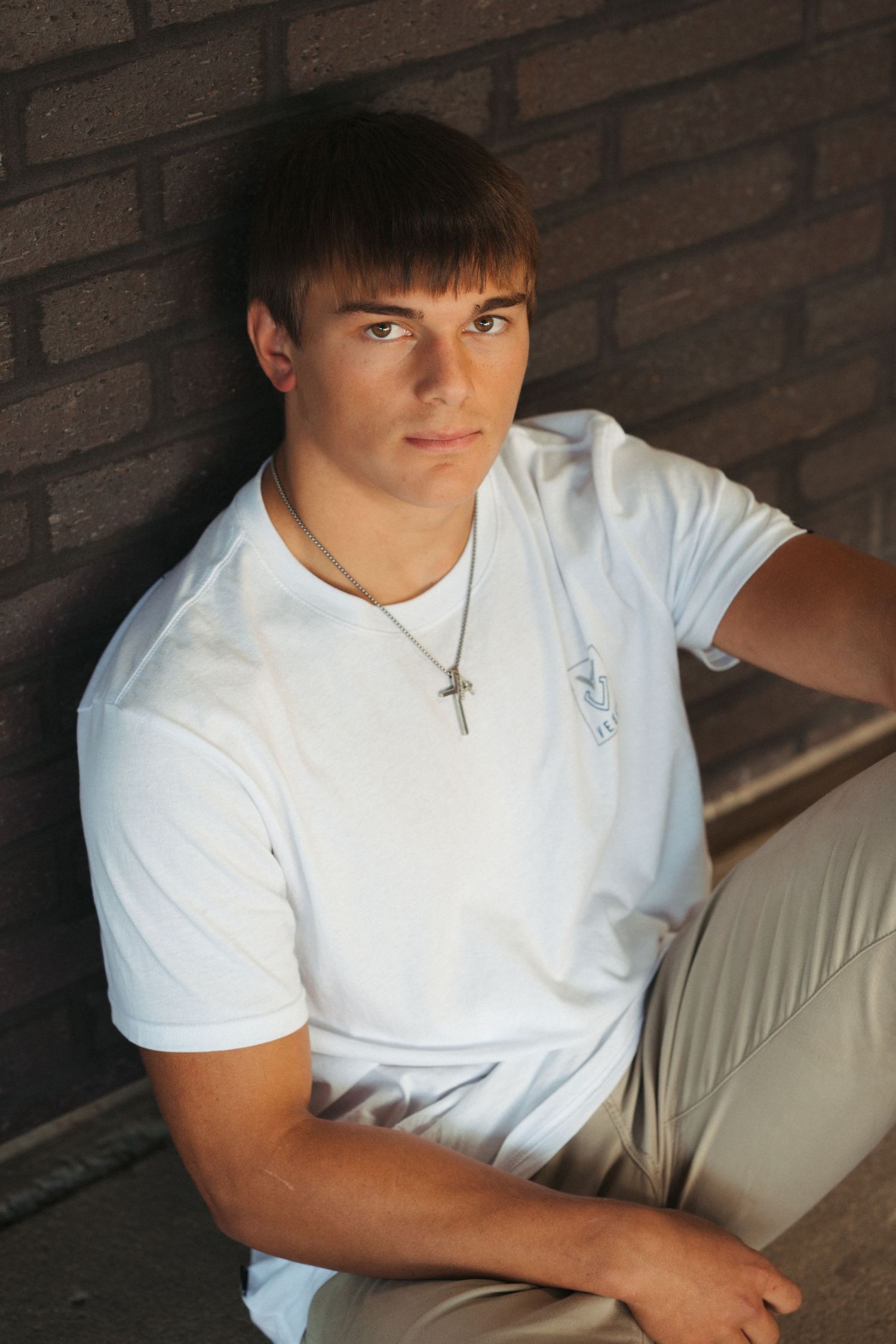 Senior boy in a white shirt photographed in an urban brick setting with clean masculine posing and modern senior portrait edits.