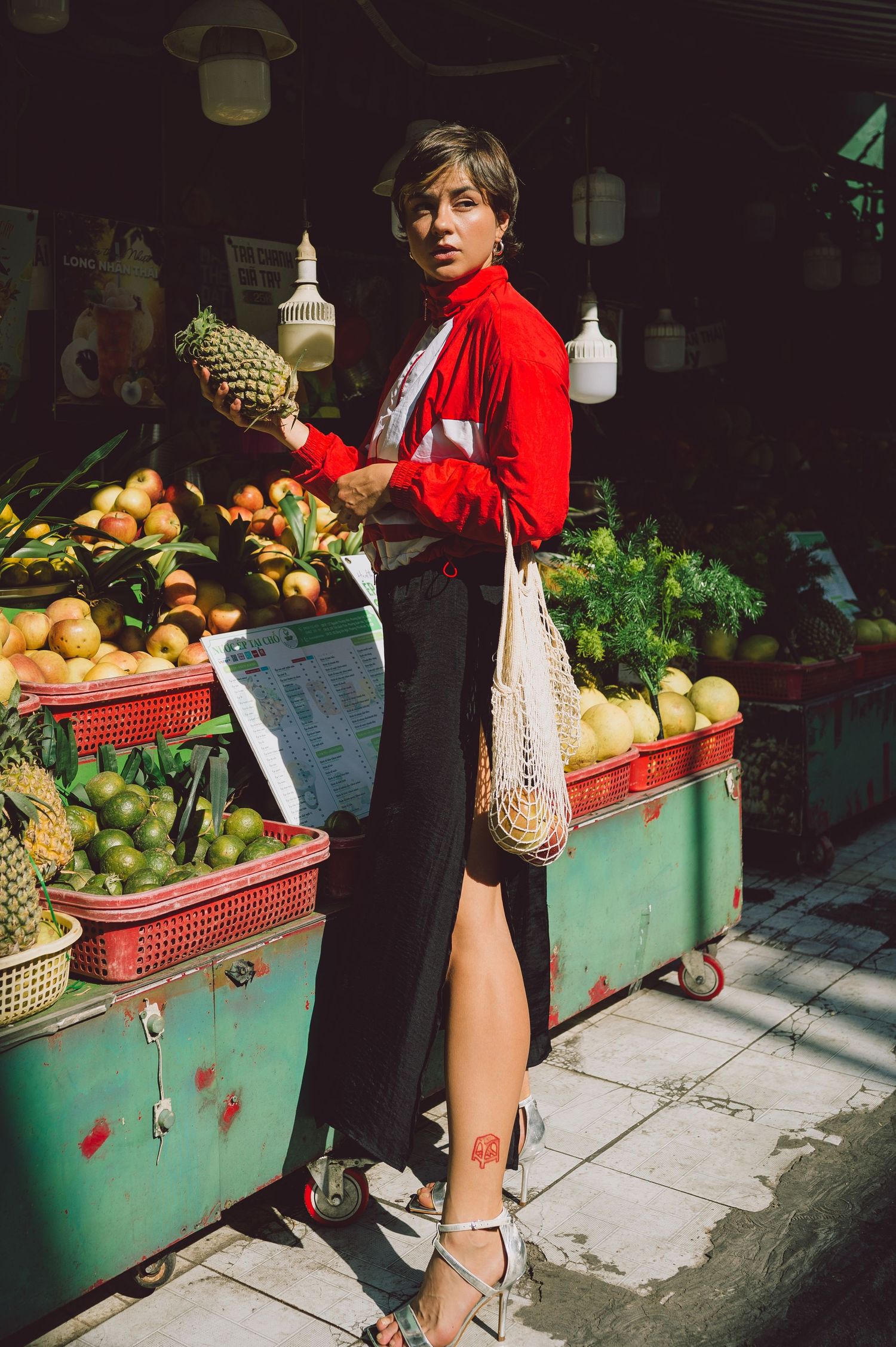 Model in a red bomber jacket carrying a mesh bag of fruits in Saigon — street editorial photography.