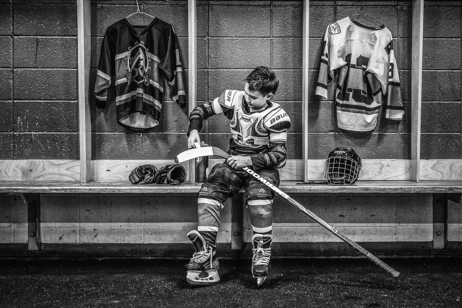 Black and white photo of hockey player sitting alone in locker room with equipment.