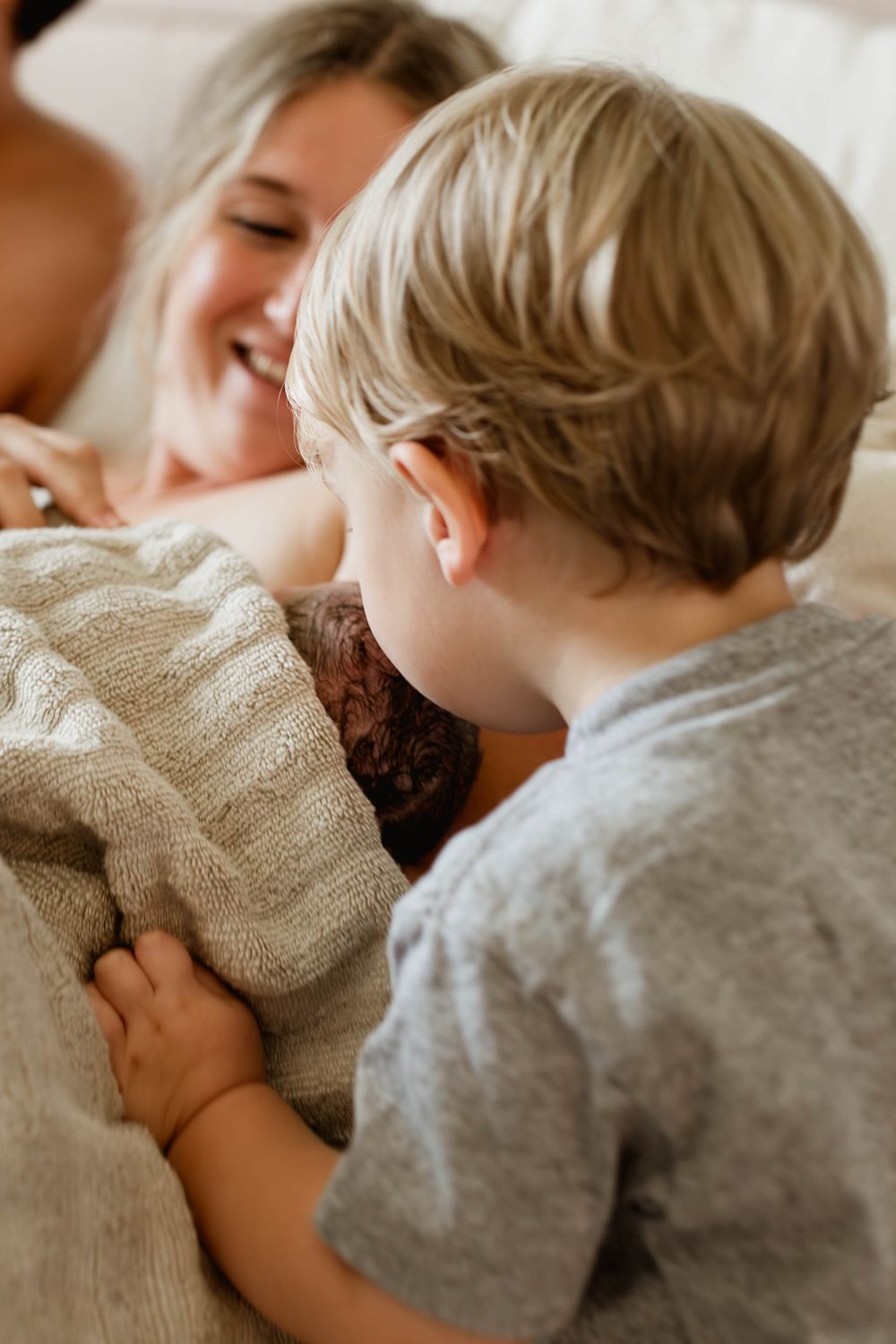 Smiling mother lying down as toddler son lovingly looks at their new baby sibling.