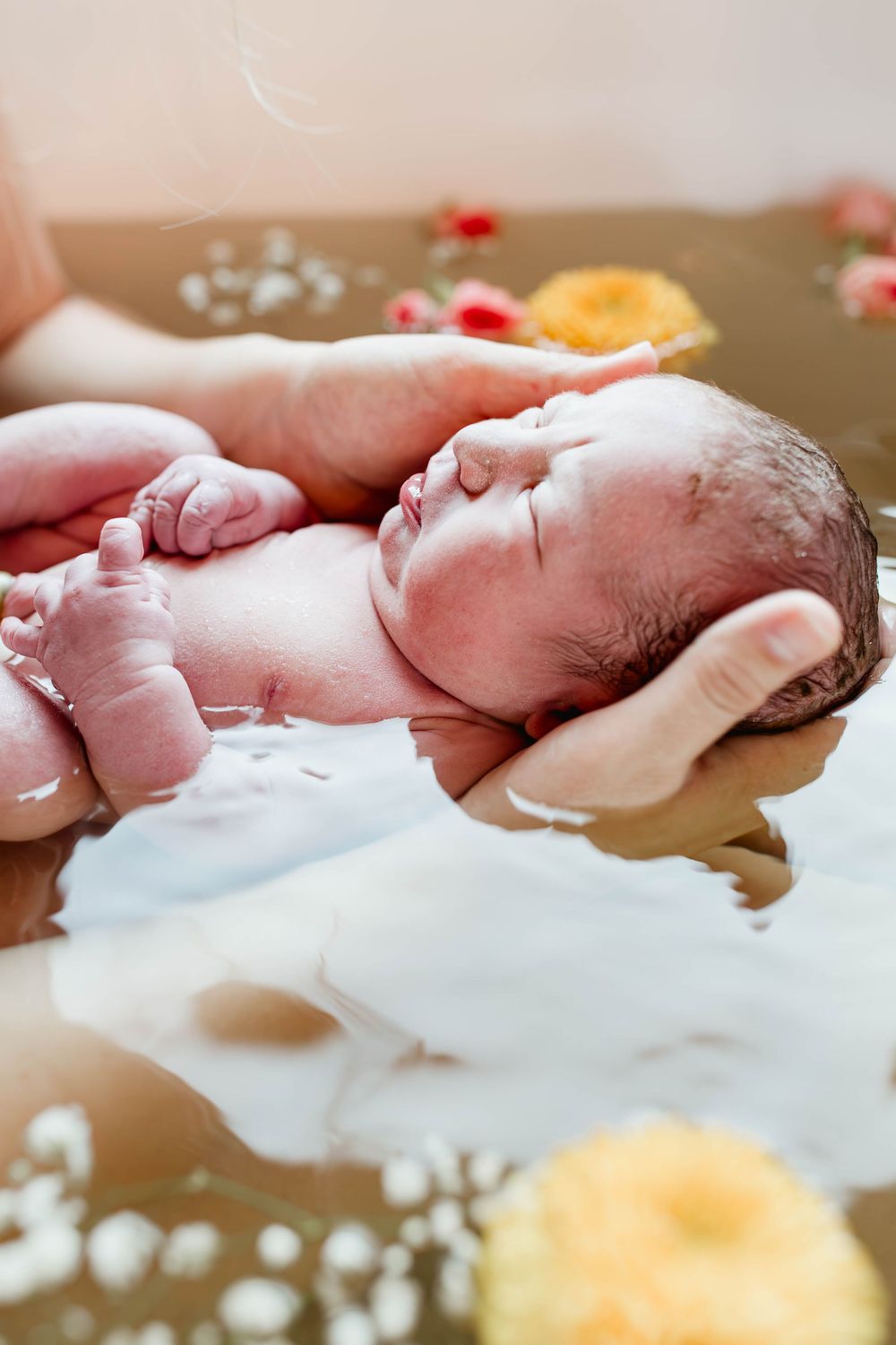 Peaceful newborn baby floating in water surrounded by flower petals during herbal bath.