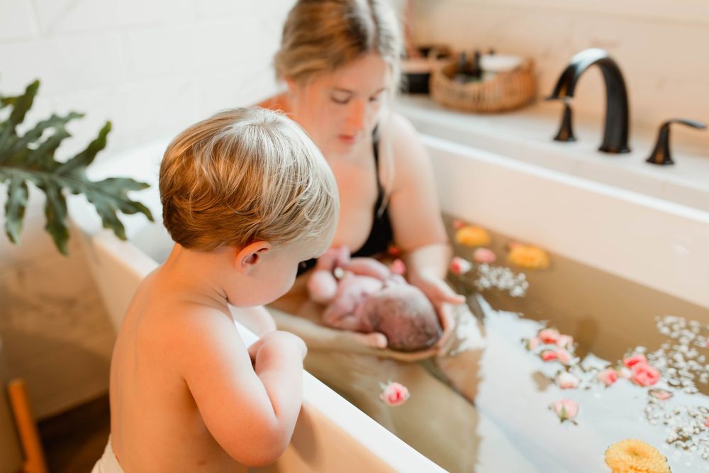 Mother and toddler during a peaceful newborn milk bath photoshoot in a white bathtub with flowers.