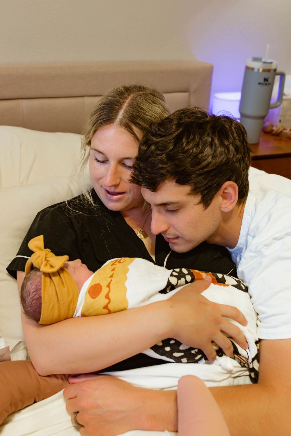 New parents smiling and cuddling their swaddled newborn baby together on a bed.