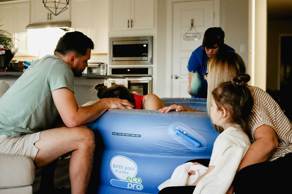 Family gathered around a blue inflatable birth pool during a home water birth in a kitchen.