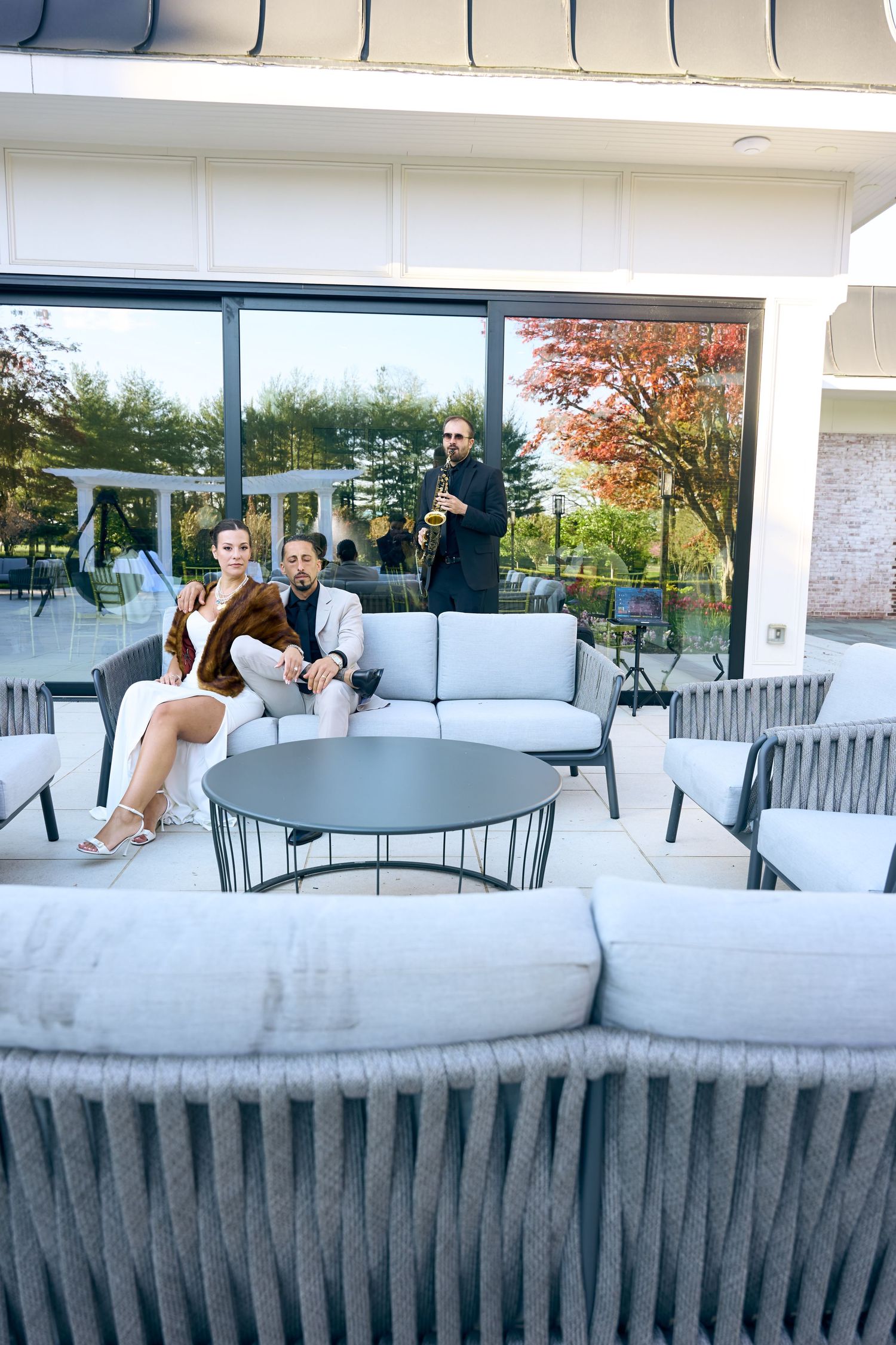 Elegant couple posing outdoors at upscale venue, woman in white dress with fur wrap, man in beige suit.
