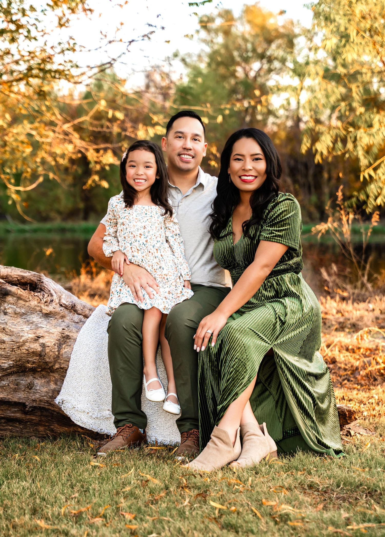 Family of three posing outdoors on a log in autumn, wearing green and floral outfits, surrounded by fall foliage.