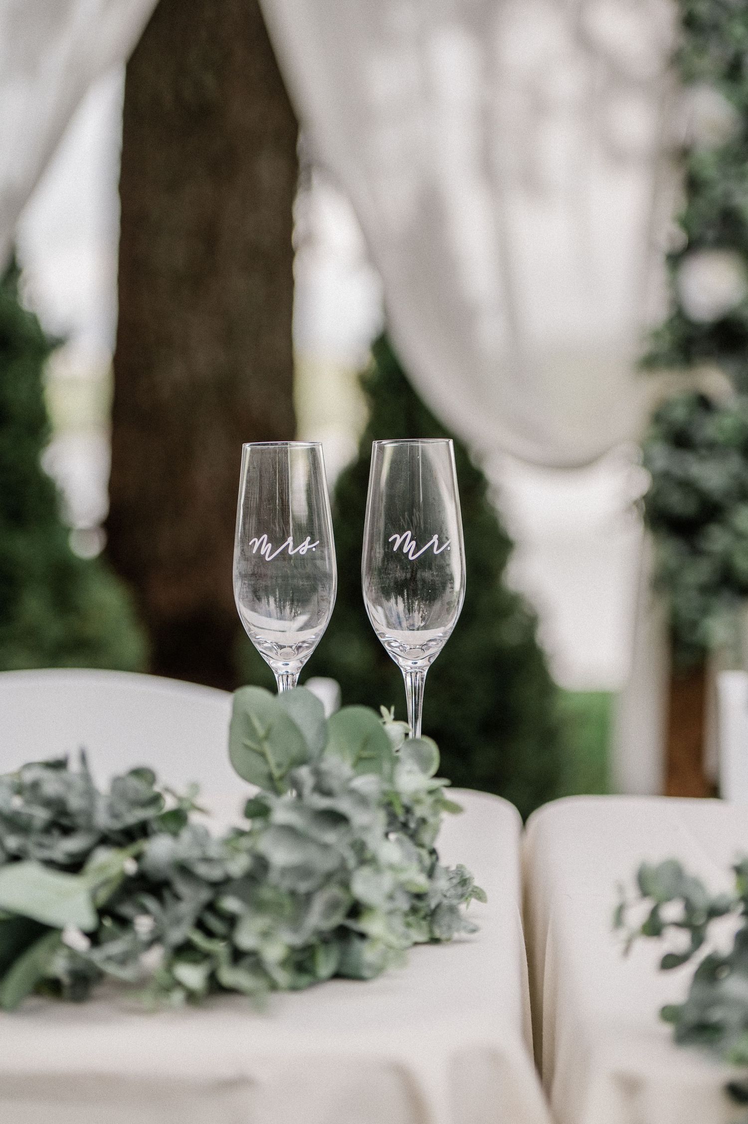 Two elegant champagne flutes engraved with Mr and Mrs at a wedding reception table with greenery.