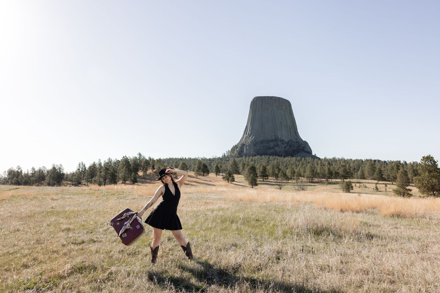 Woman in black dress carrying suitcase walks through meadow with Devils Tower monument in background.