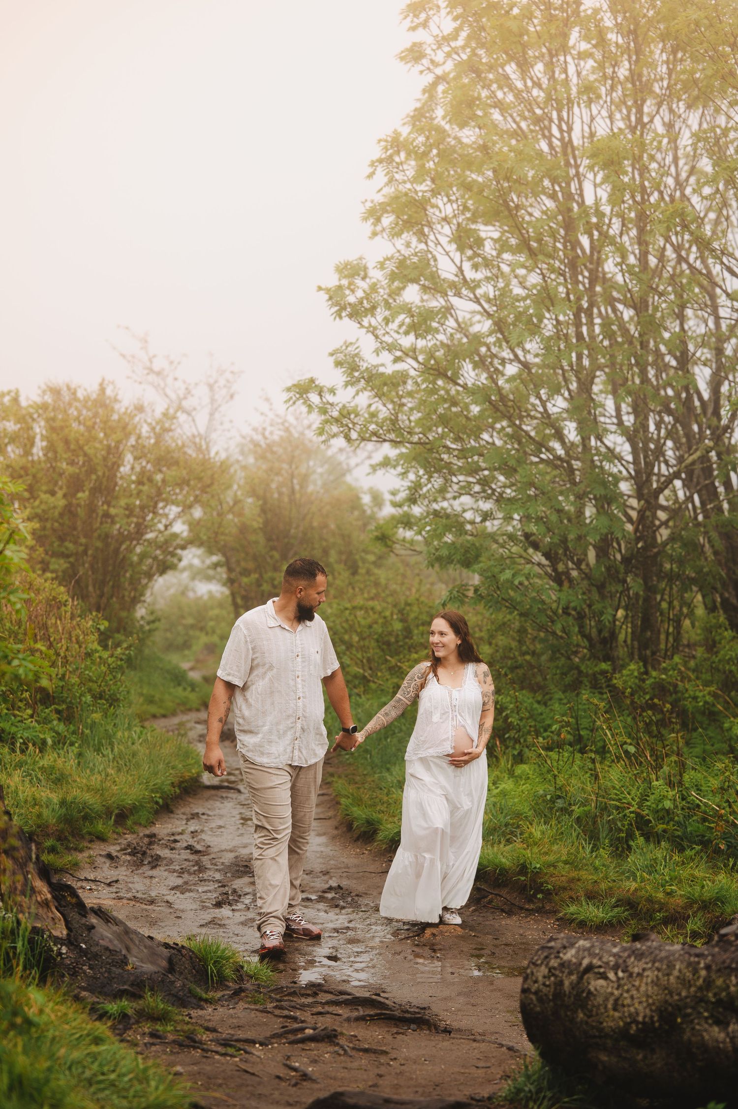 Pregnant couple holding hands walking on a misty forest path, woman in white dress, man in casual attire.