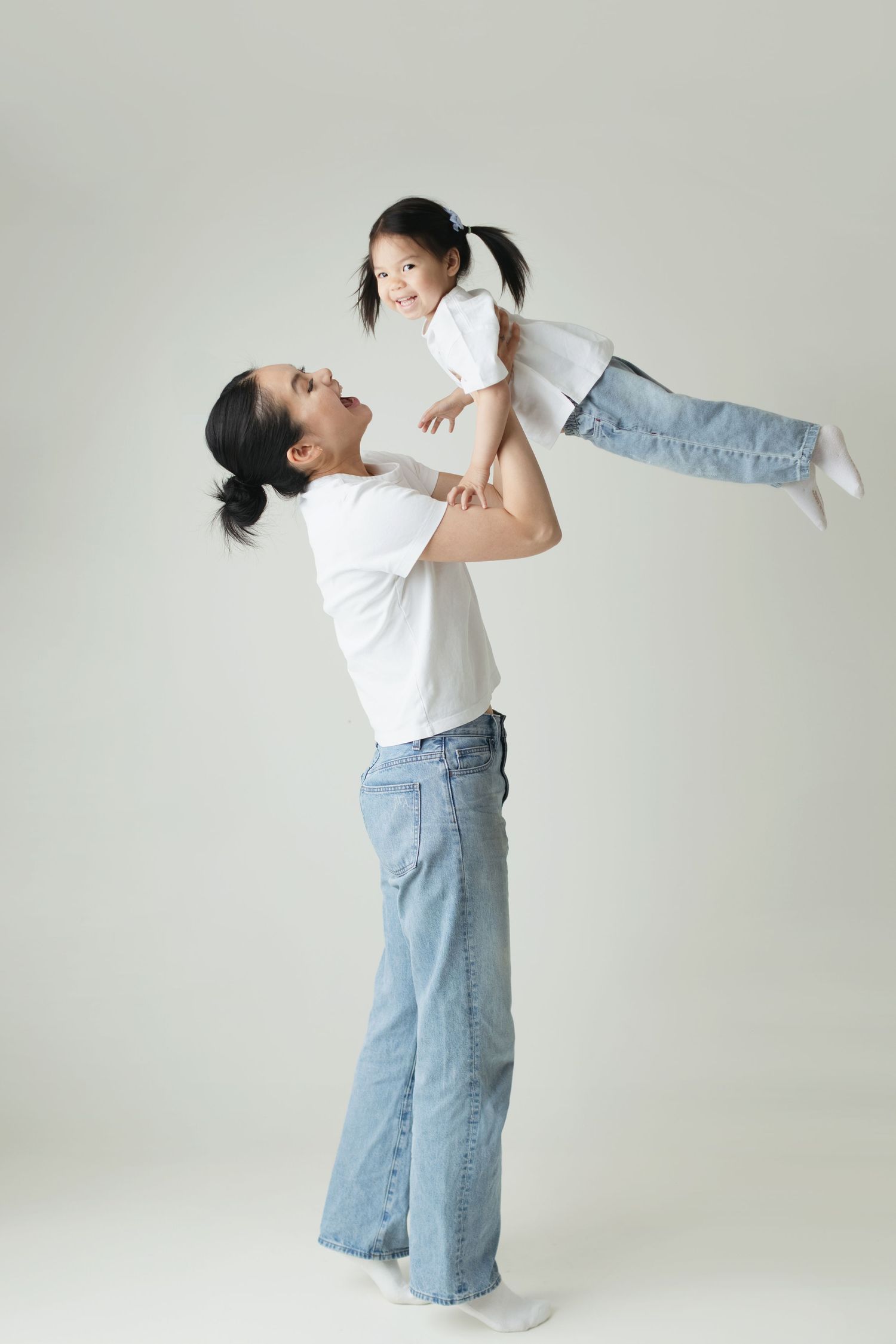 A mother joyfully lifts her young daughter in the air, both wearing white shirts and jeans on a light background.