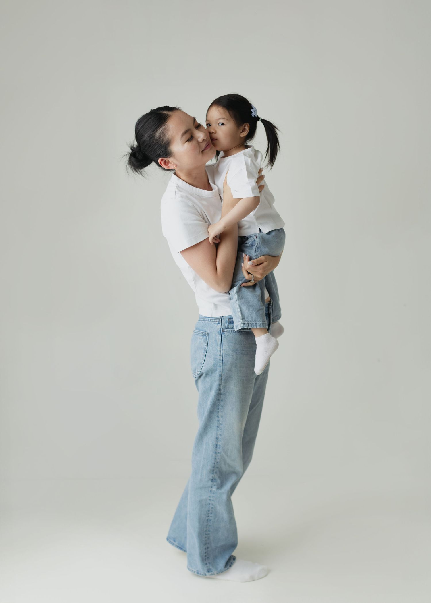 A woman in white tee and jeans holds and kisses a young girl in a white top and denim skirt.