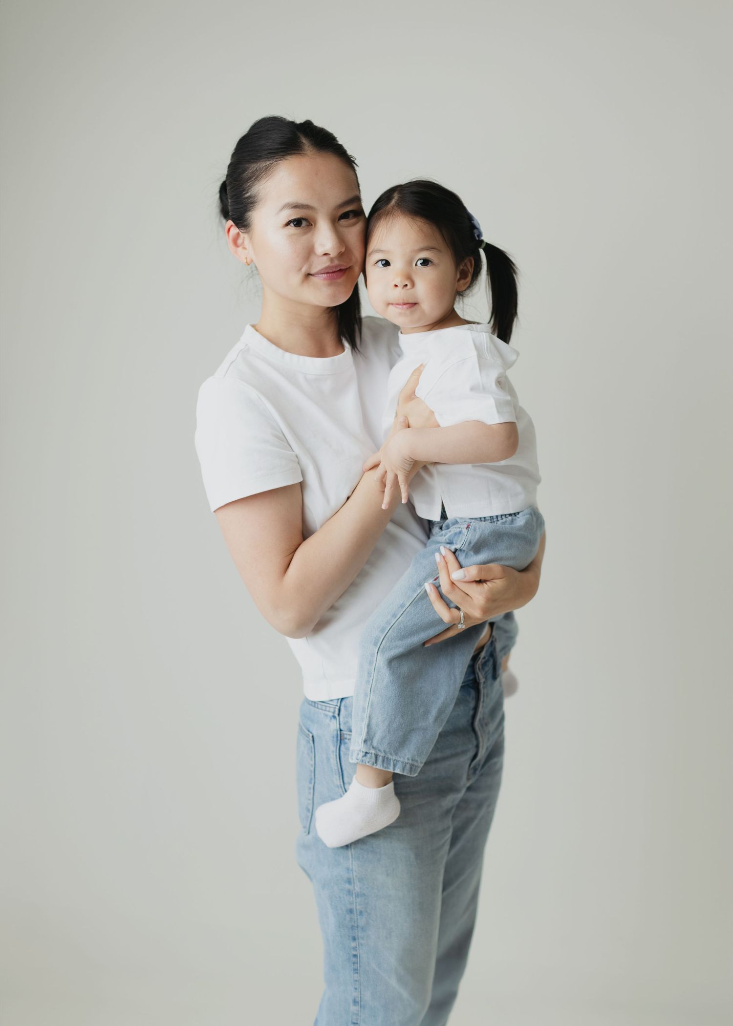 A woman in a white t-shirt and jeans holds a young toddler girl in matching white top against a light background.