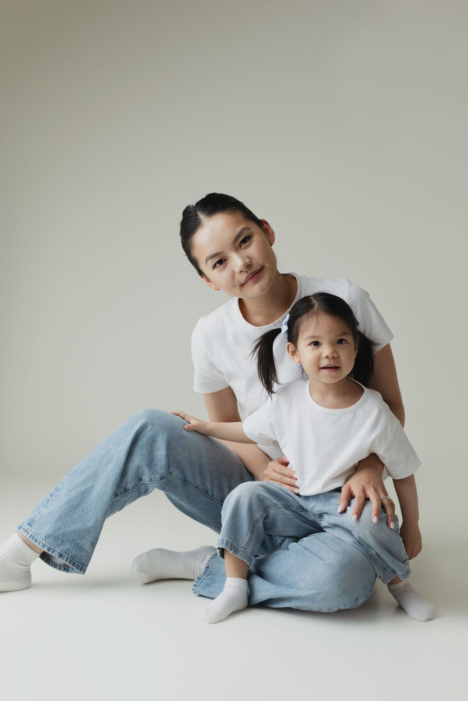 Mother and daughter in matching white tees and jeans pose together on white background.