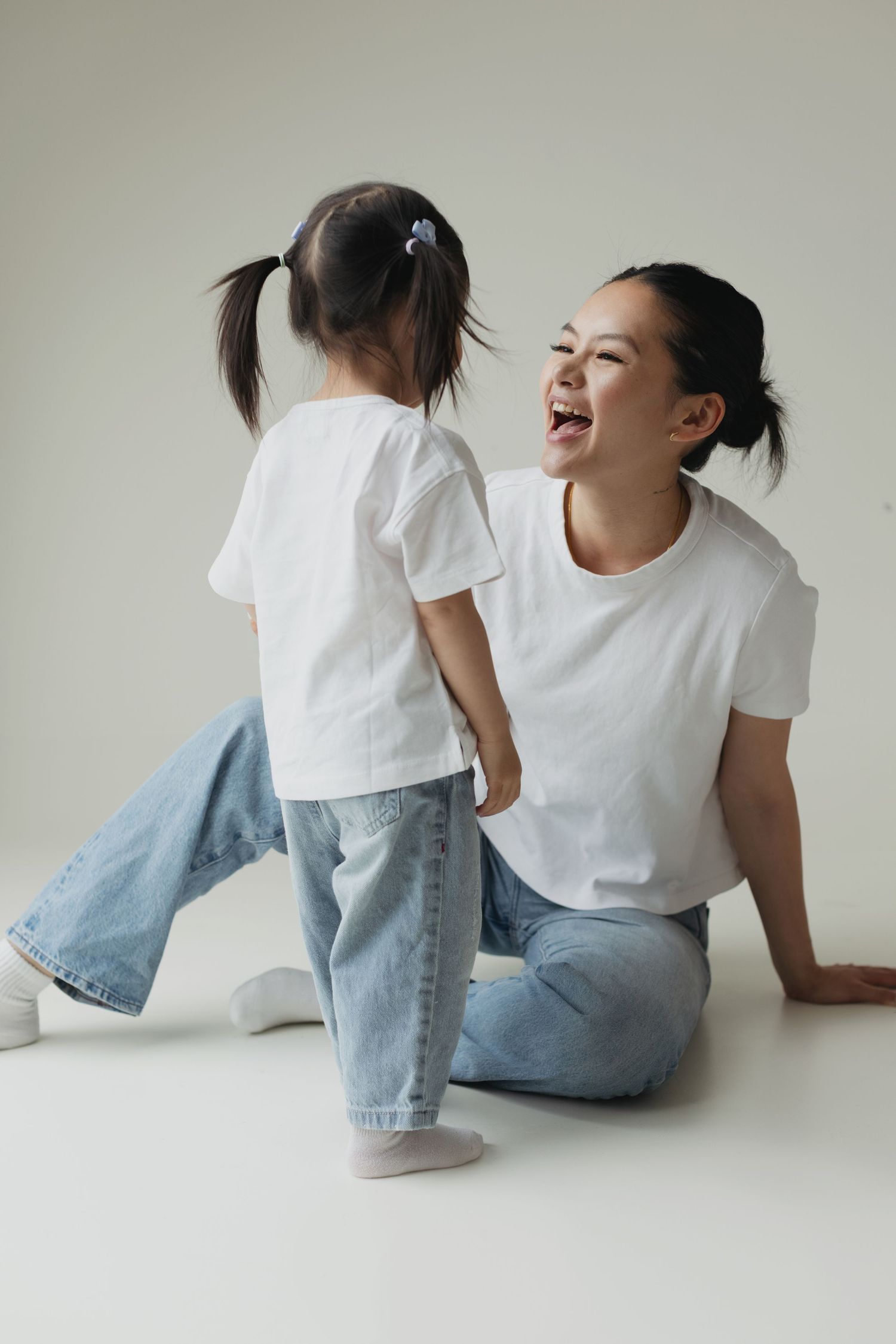 Laughing mother in white tee and jeans plays with young daughter in studio setting.