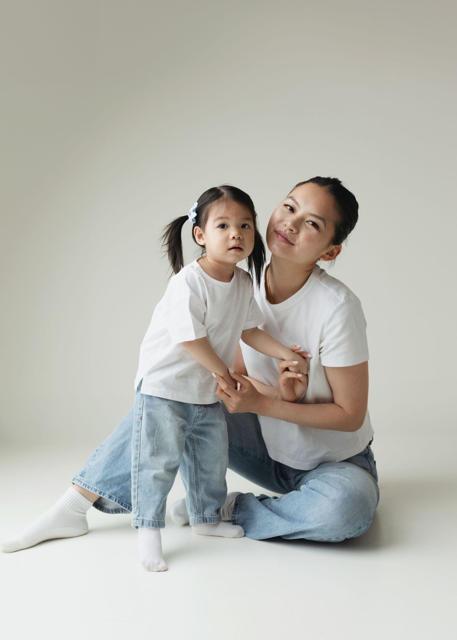 A woman and young girl in matching white t-shirts and jeans sit together on a white background.