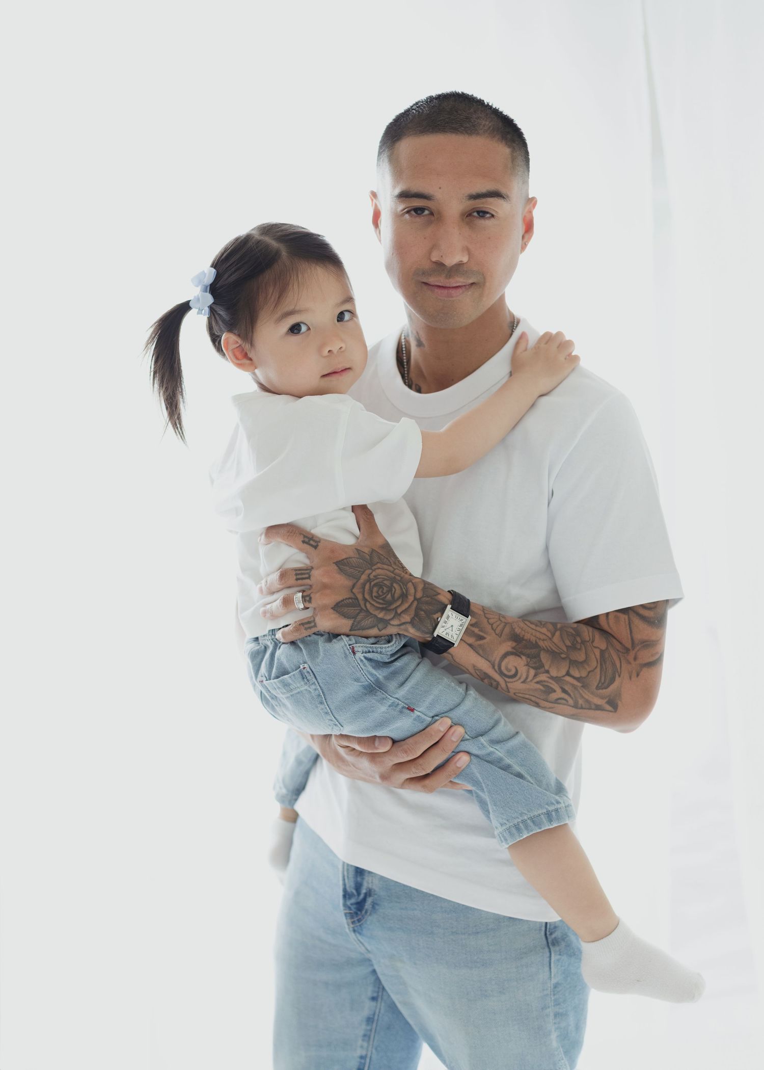 Man with tattoos in white t-shirt and jeans holds a young girl with pigtails wearing a white dress.
