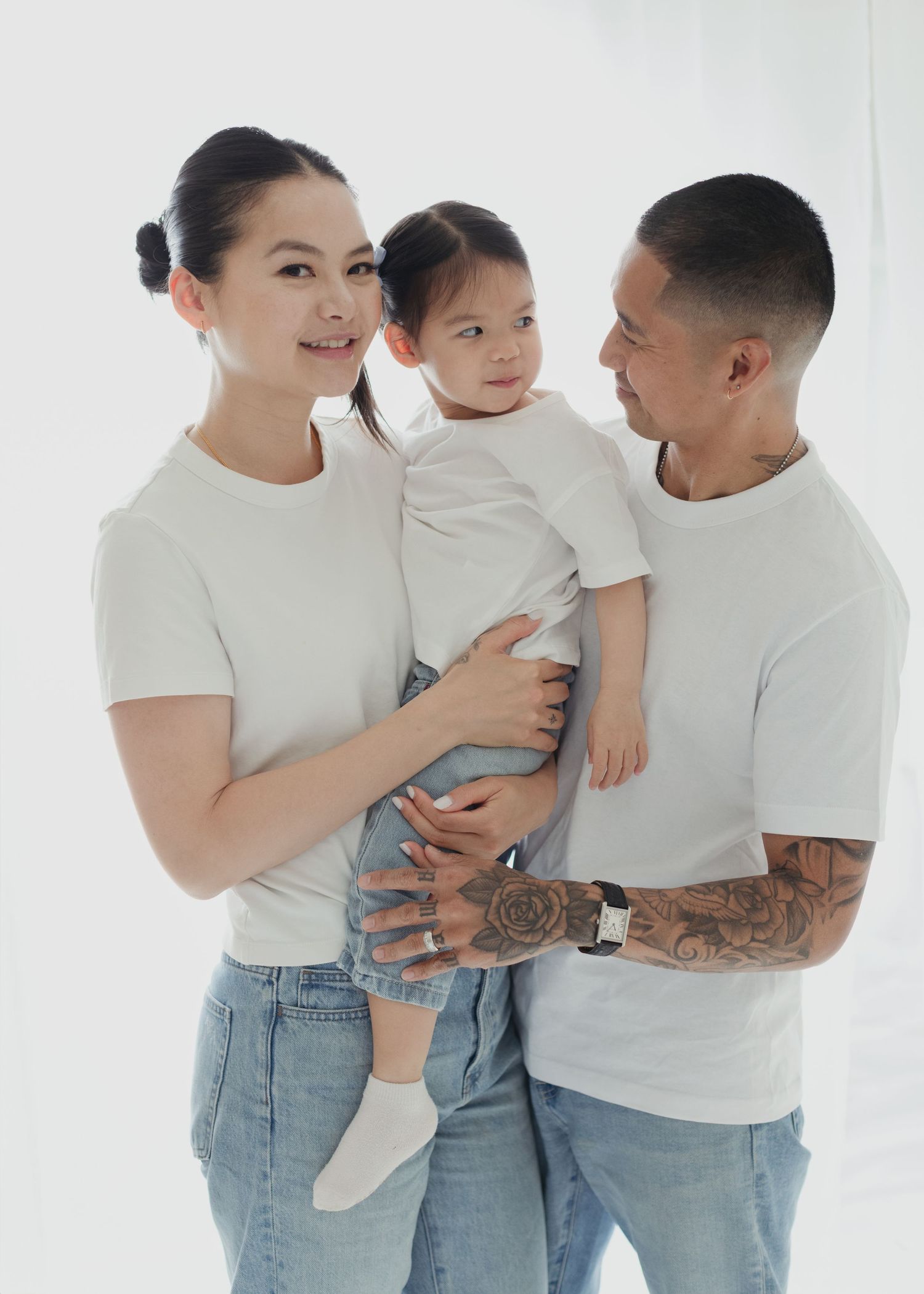 A happy family of three wearing matching white t-shirts and jeans, with tattooed father holding their toddler beside smiling mother.