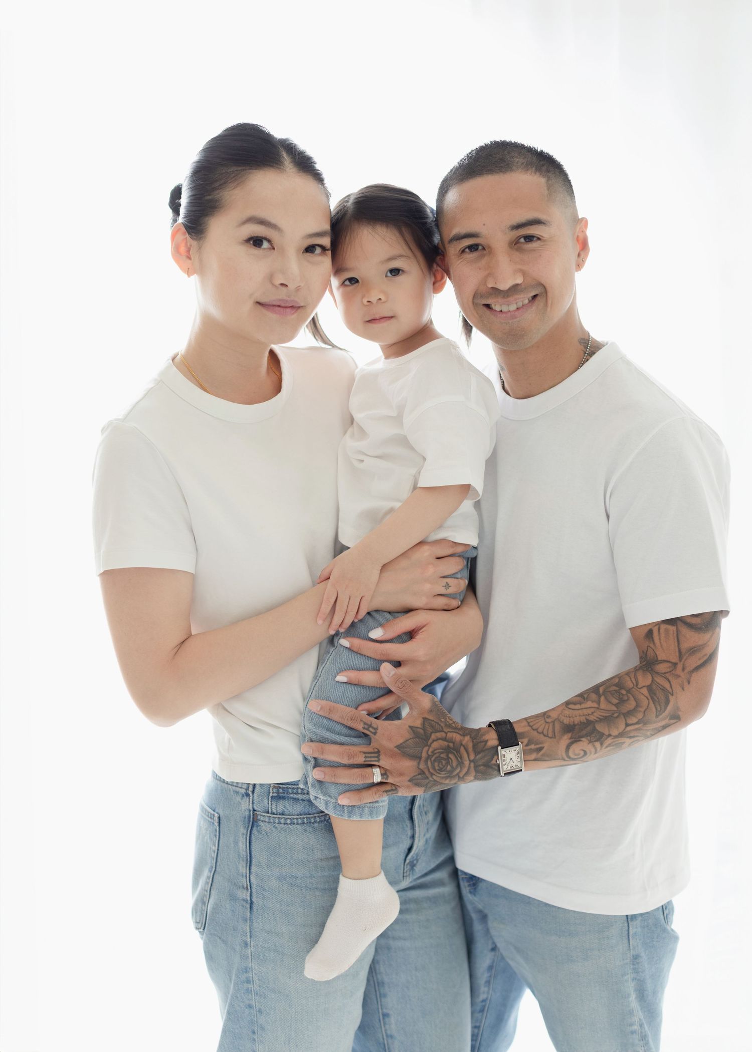 A smiling family of three in matching white tees and jeans, parents holding their toddler daughter against a white background.