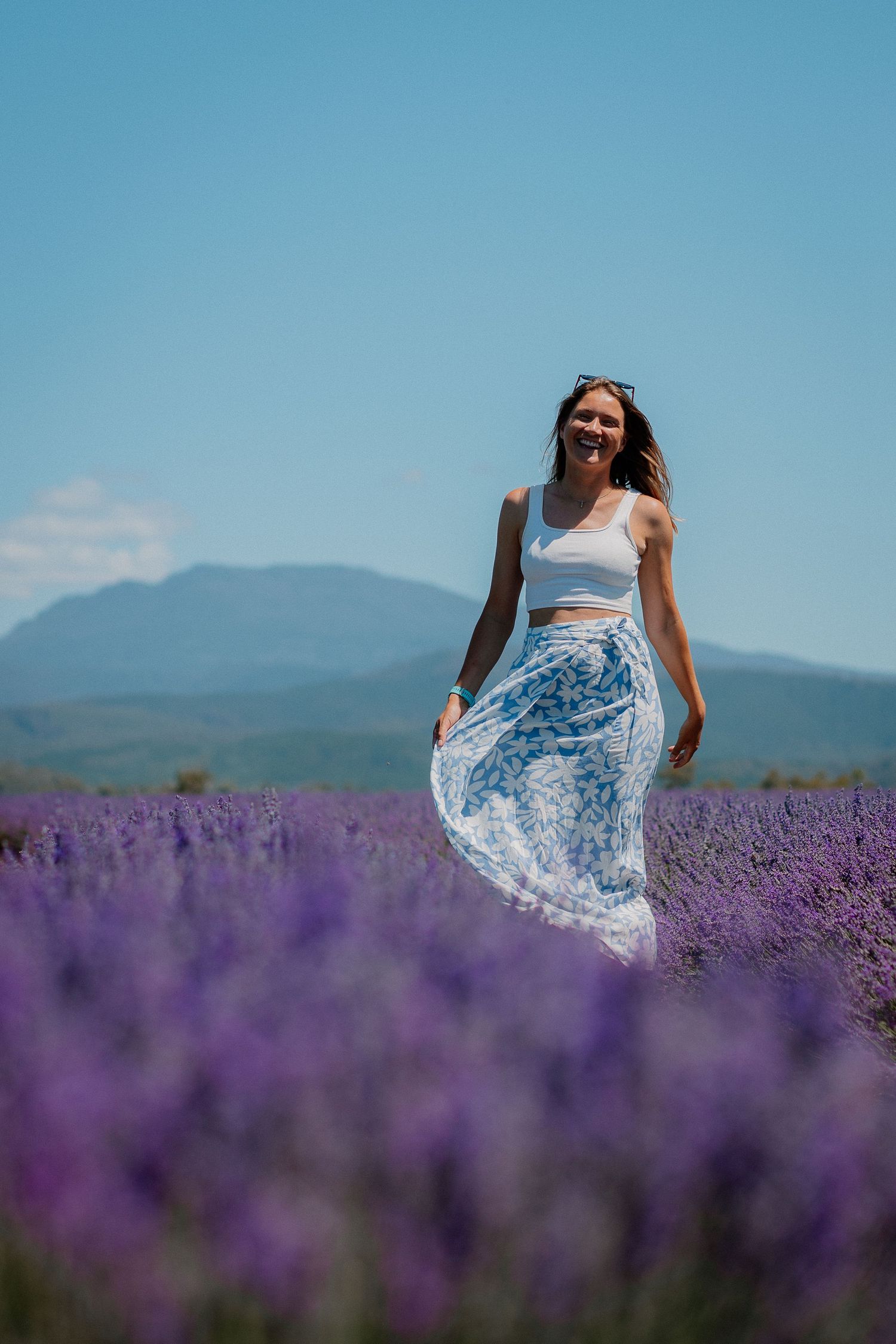 Smiling woman in white crop top and floral skirt walking through purple lavender field with mountains in background.
