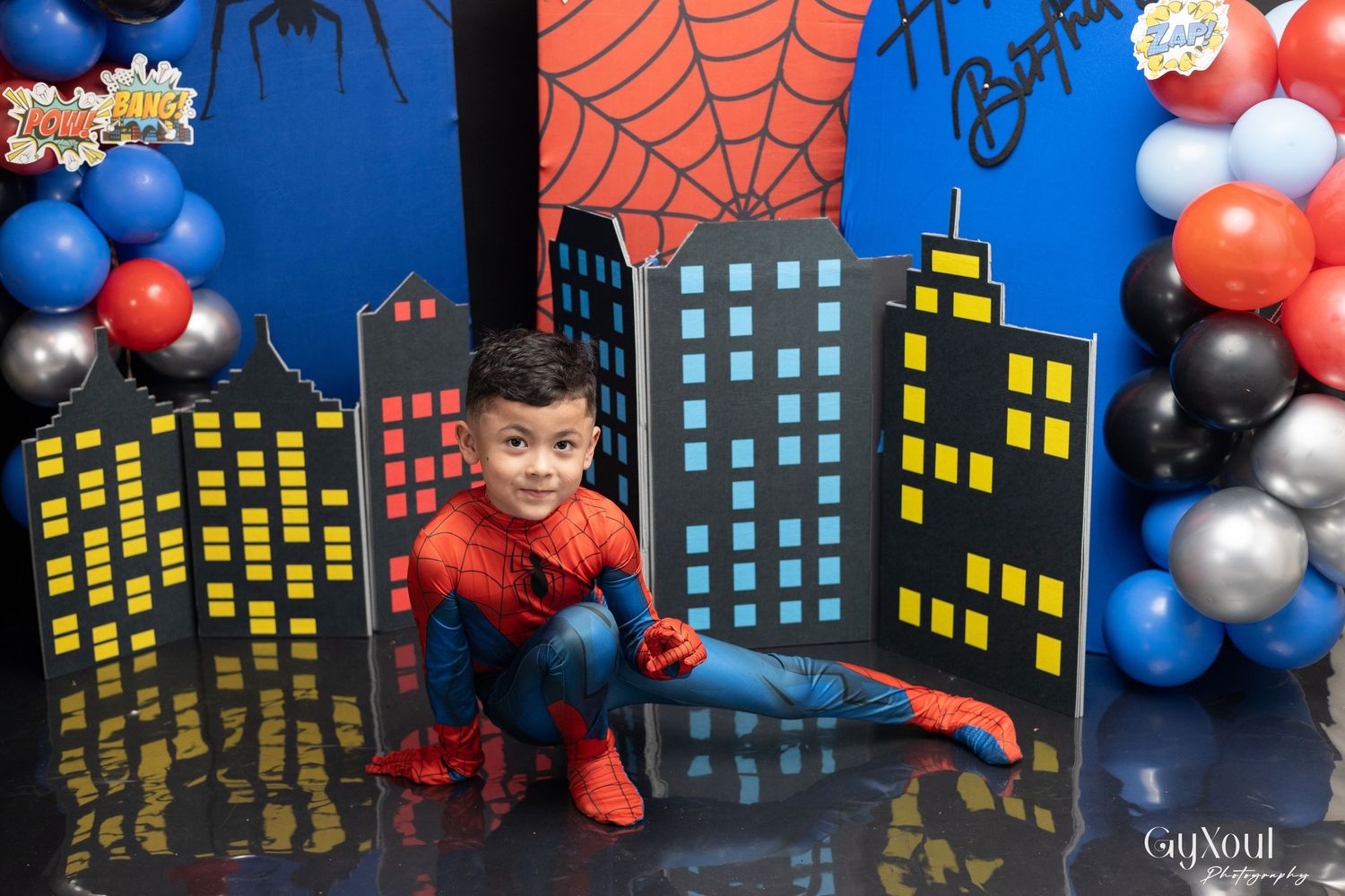 Boy in Spiderman costume poses at a superhero-themed birthday party with colorful balloons and city backdrop.