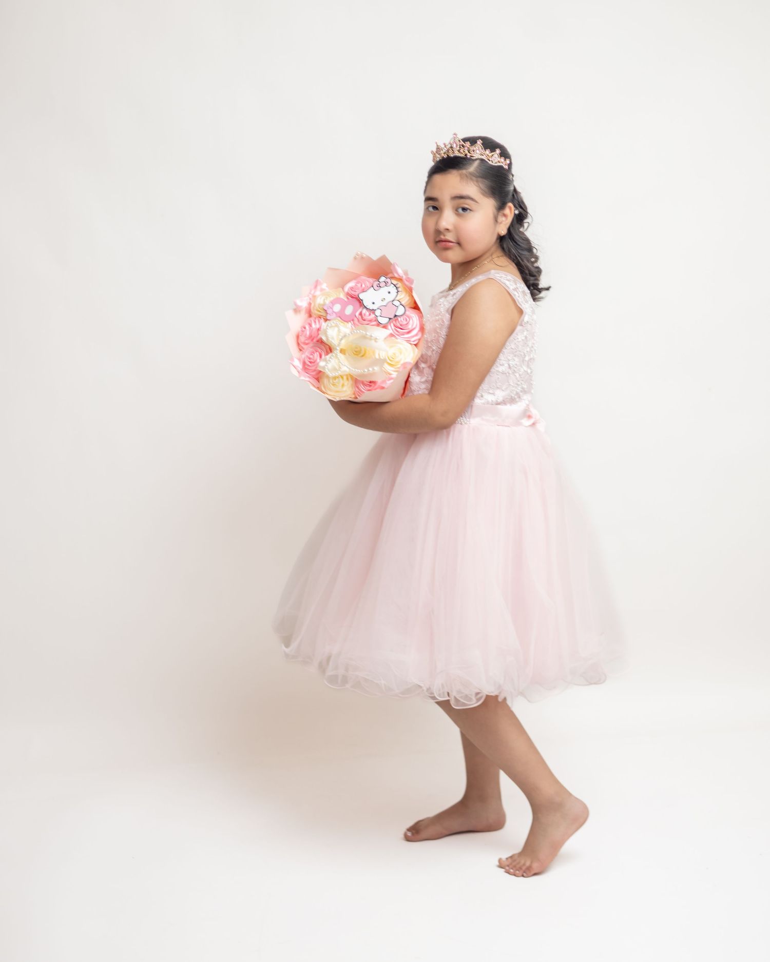Young girl in pink tulle dress and tiara holds floral balloon bouquet, posing barefoot against white background.