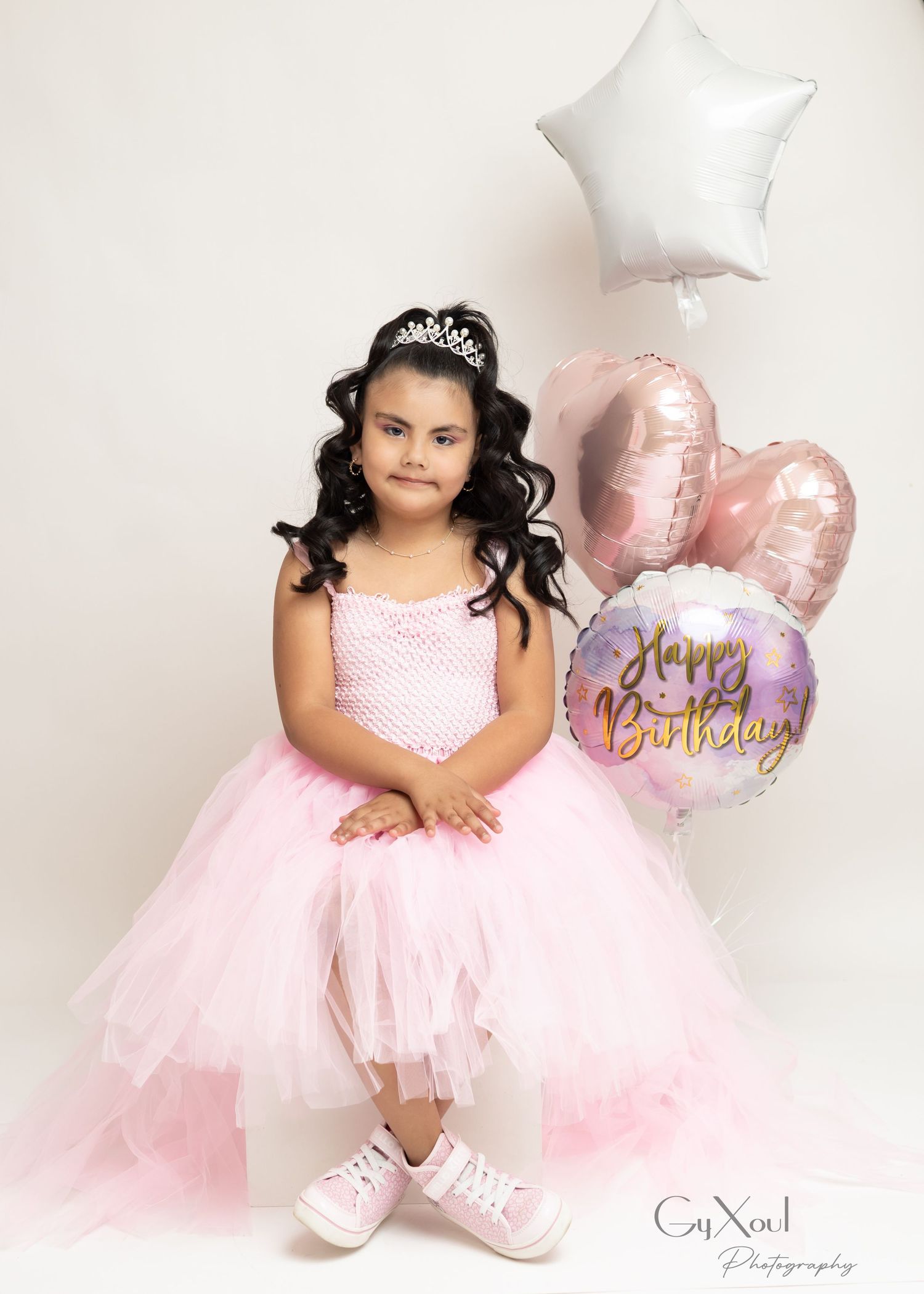 Young girl in pink tutu dress and tiara poses with Happy Birthday balloons for a birthday photo session.