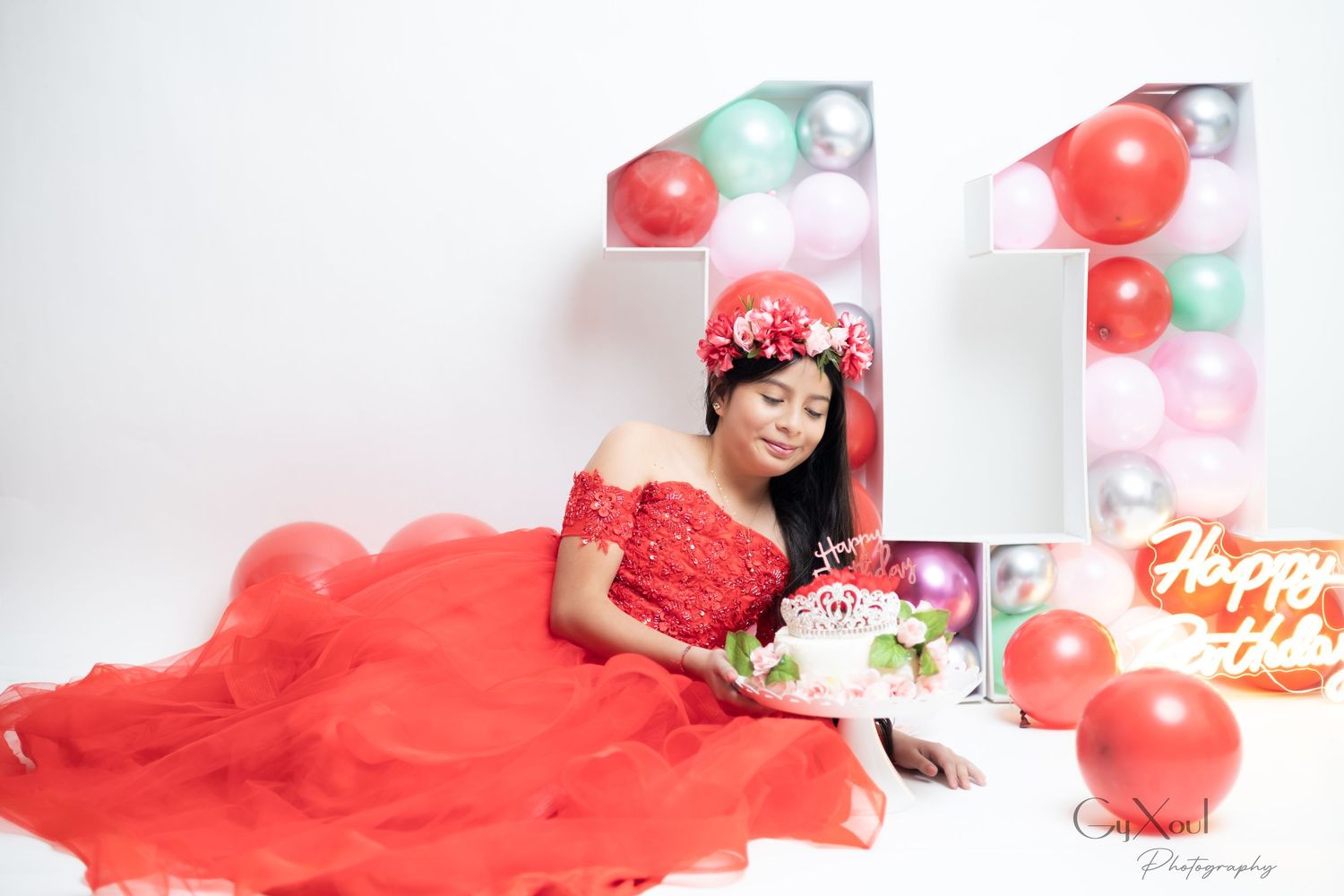 Young woman in red off-shoulder gown sits beside number 11 balloon display with birthday cake and Happy Birthday sign.