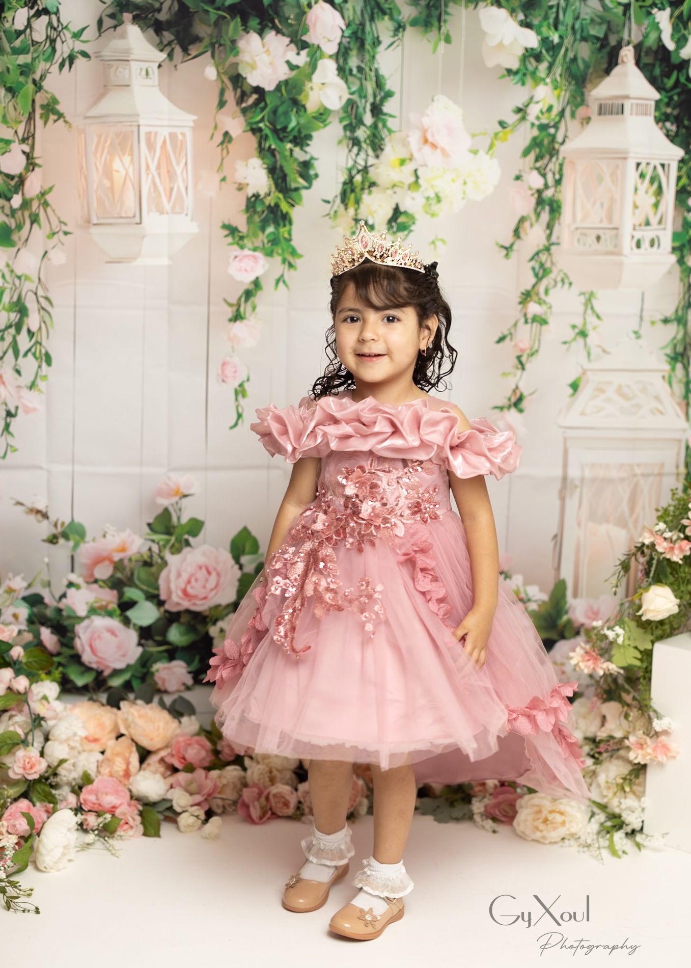 Young girl in pink floral dress and crown poses among roses and lanterns in a dreamy birthday portrait.