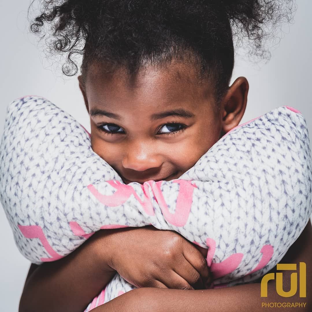 Adorable young Black girl with curly hair smiles while hugging a decorative pillow in studio.